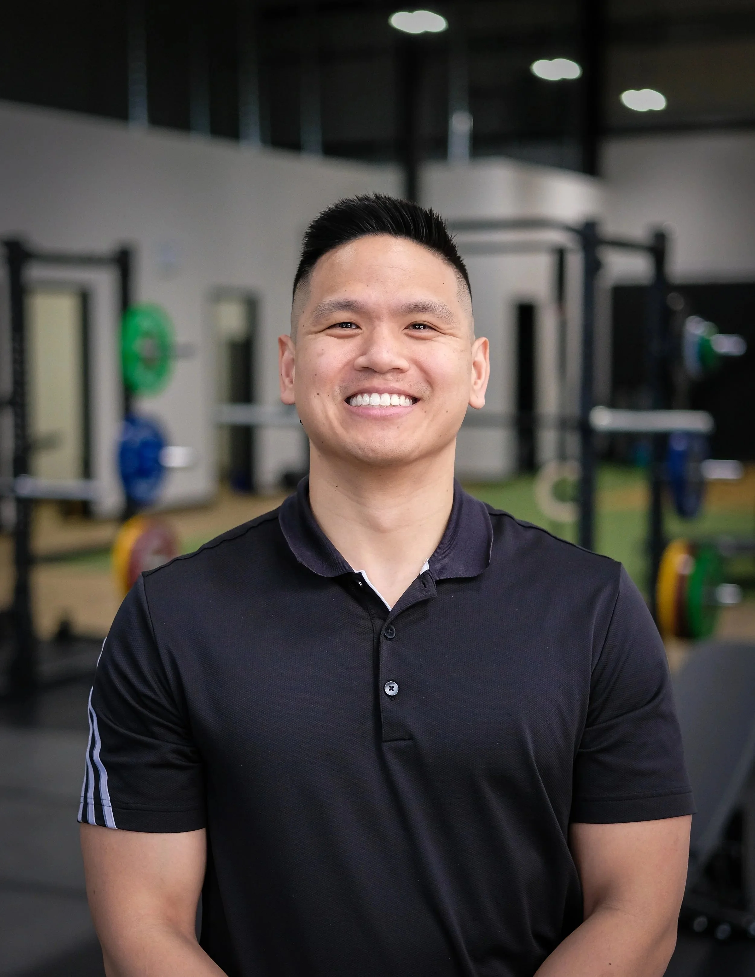 A male physiotherapist with short dark hair and a broad smile, wearing a black polo shirt with white stripes on the sleeves, standing indoors in a gym space of a physical therapy clinic with workout equipment in the background.