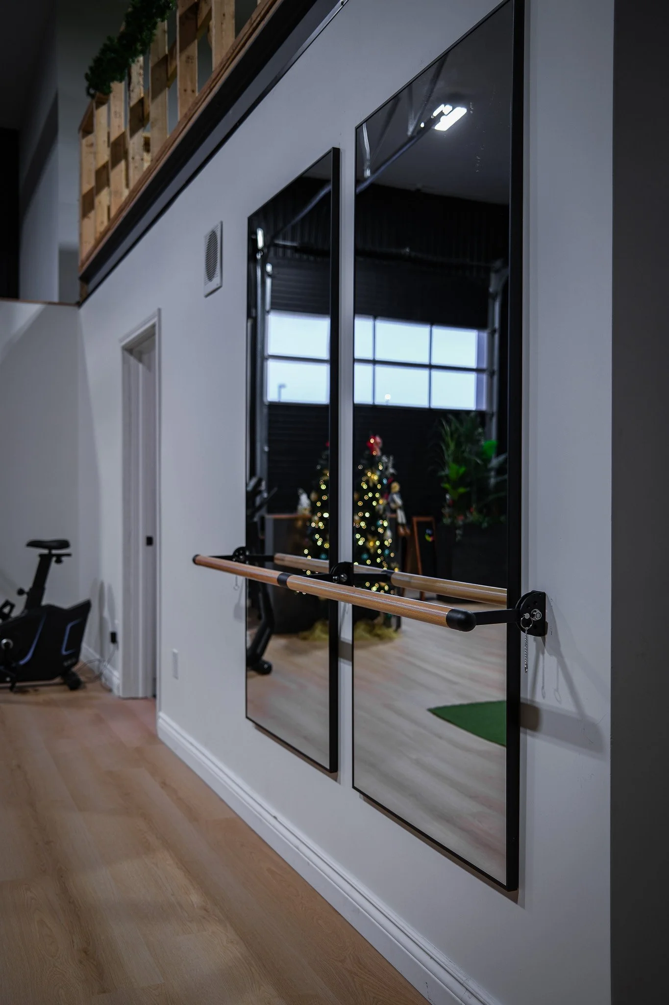 A mirror with a ballet barre reflecting a Christmas tree decorated with lights and ornaments in a room with black walls, large windows, and plants.