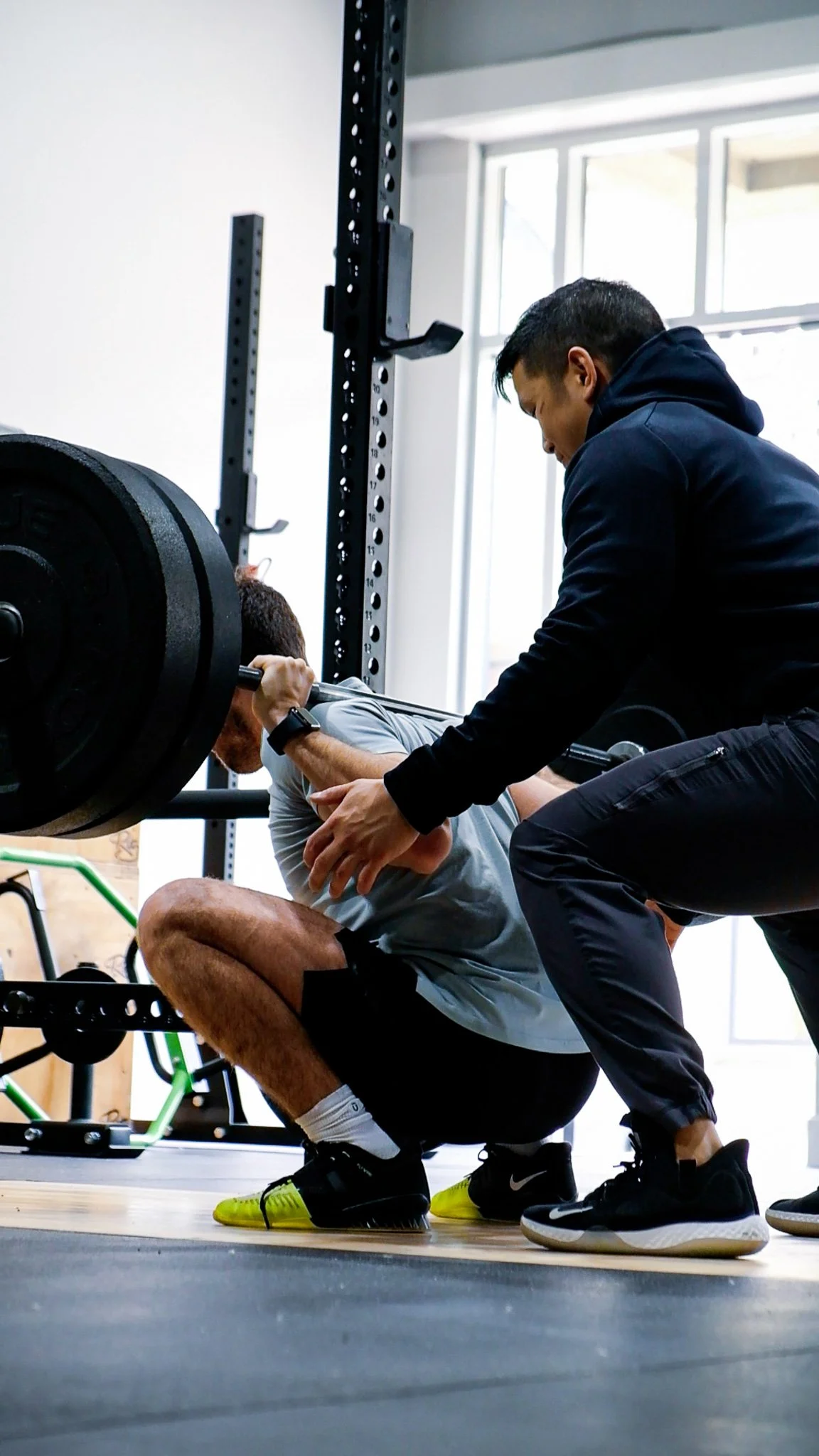 A man performing a squat exercise with a barbell and weights, supervised by a physiotherapist in a gym.