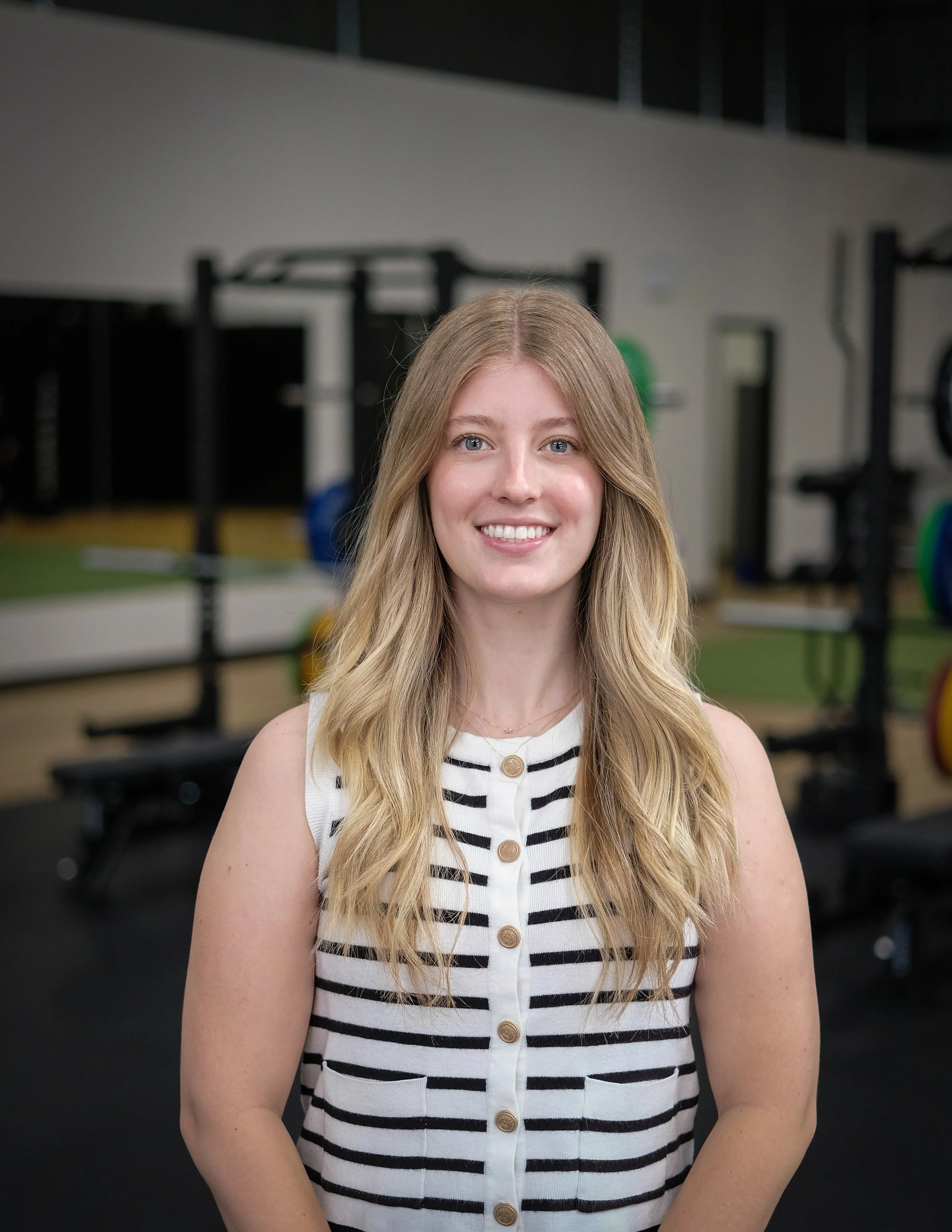 A young woman with long blonde hair and blue eyes smiling in a gym setting, wearing a sleeveless white and black striped button-up top.