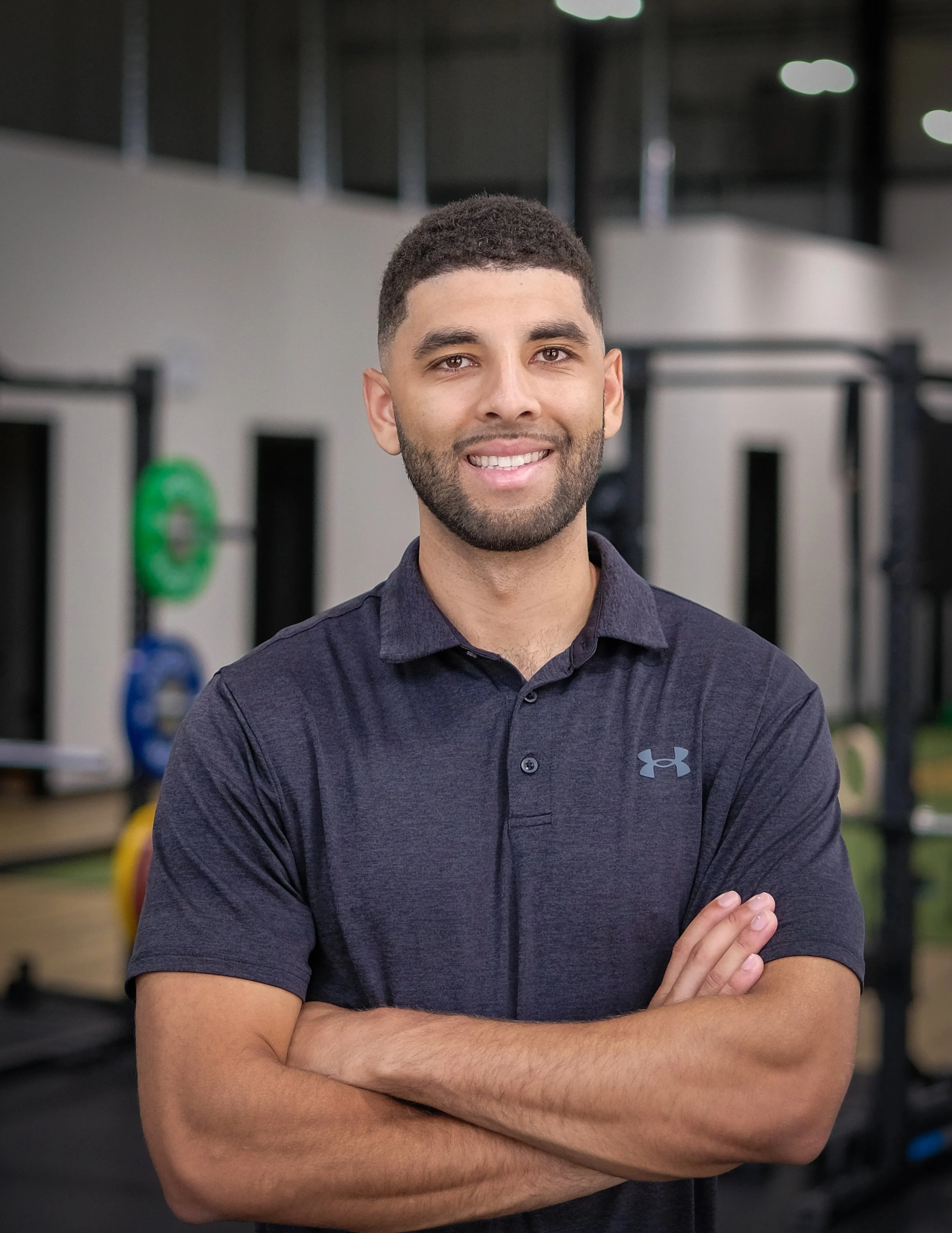 A male chiropractor with short curly hair and a beard, smiling and wearing a dark blue polo shirt, standing with arms crossed in a gym.