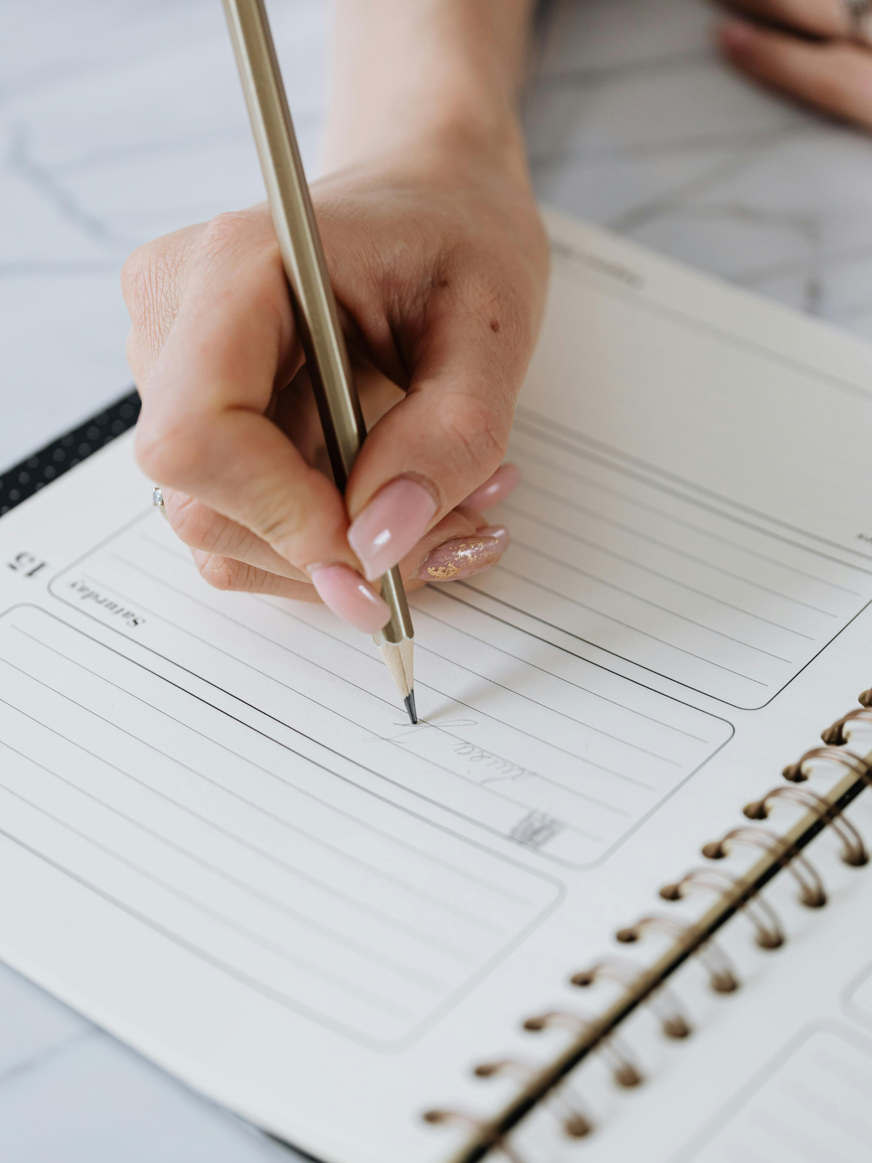 A person is writing in a planner with a gold pen, open to a page with lines and sections for notes or goals.