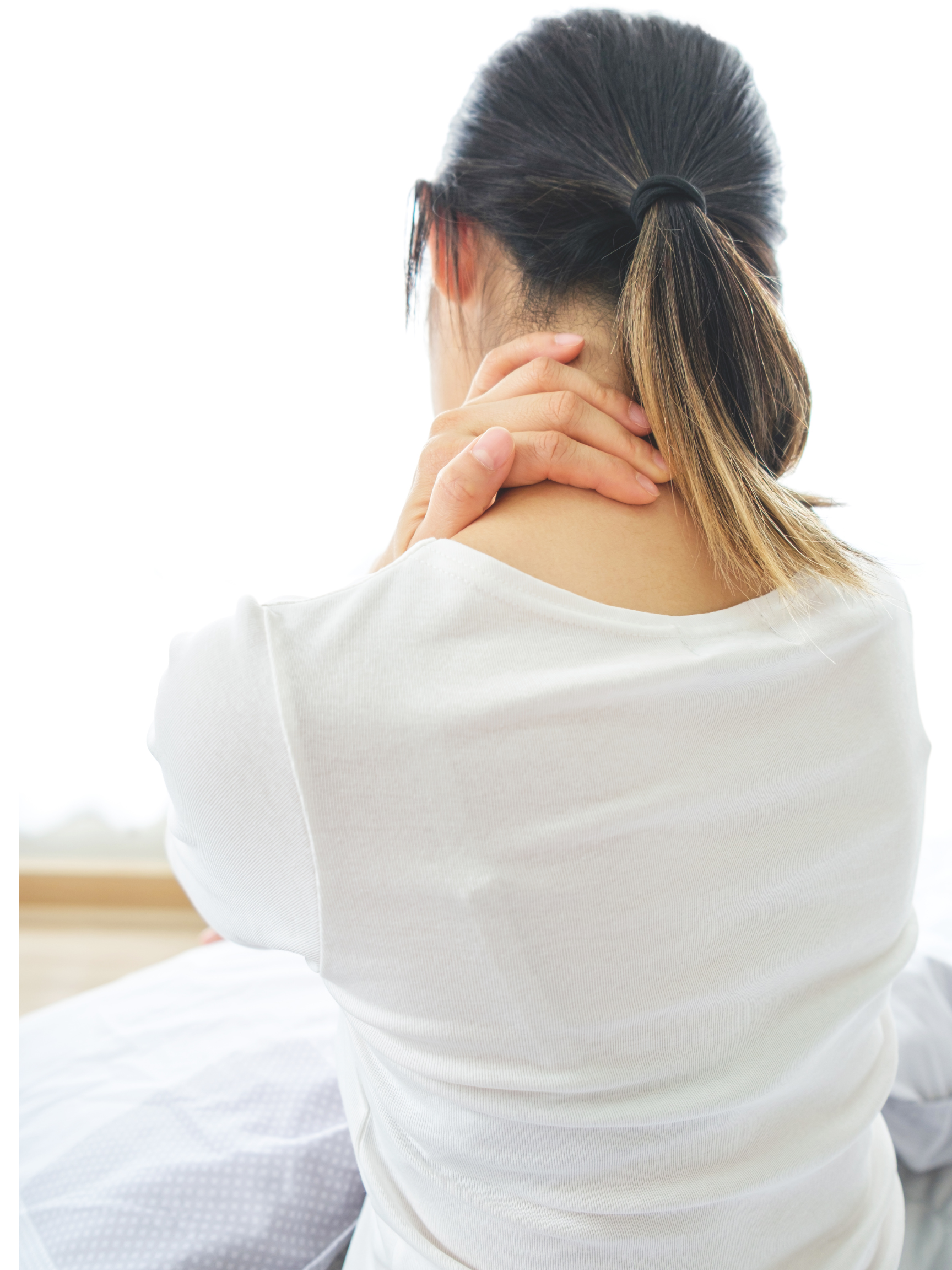 Back view of a woman with dark hair tied in a ponytail, holding her neck with her right hand, wearing a white shirt, in a bright, softly lit environment.