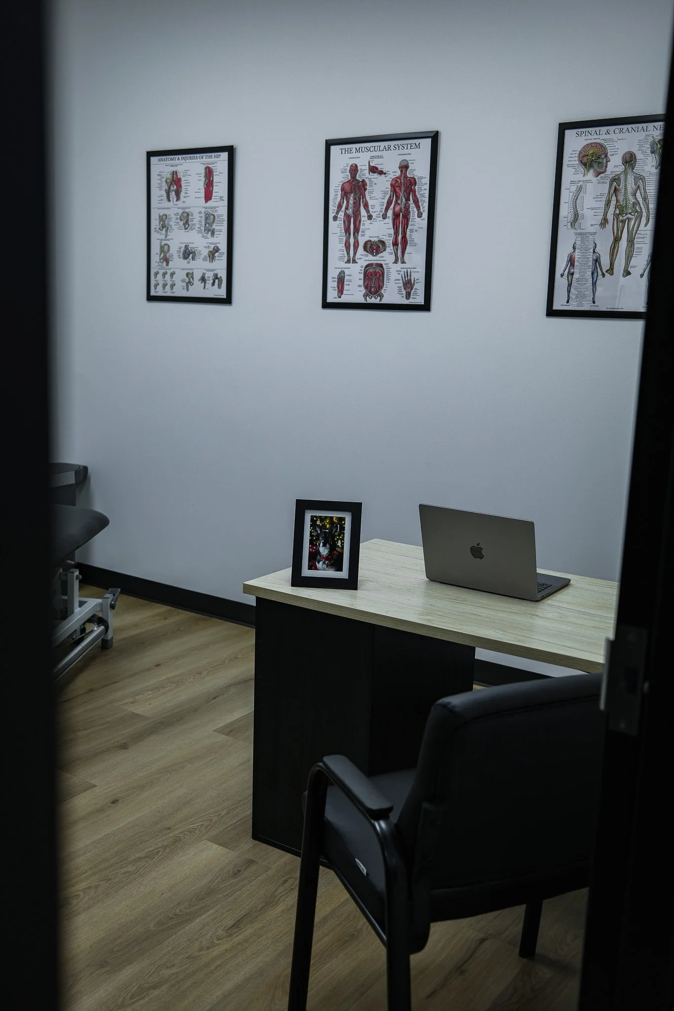 Office or waiting room with framed anatomical posters on the white wall, a small desk with a framed photo, a closed Apple MacBook, black chairs, and a wooden floor.