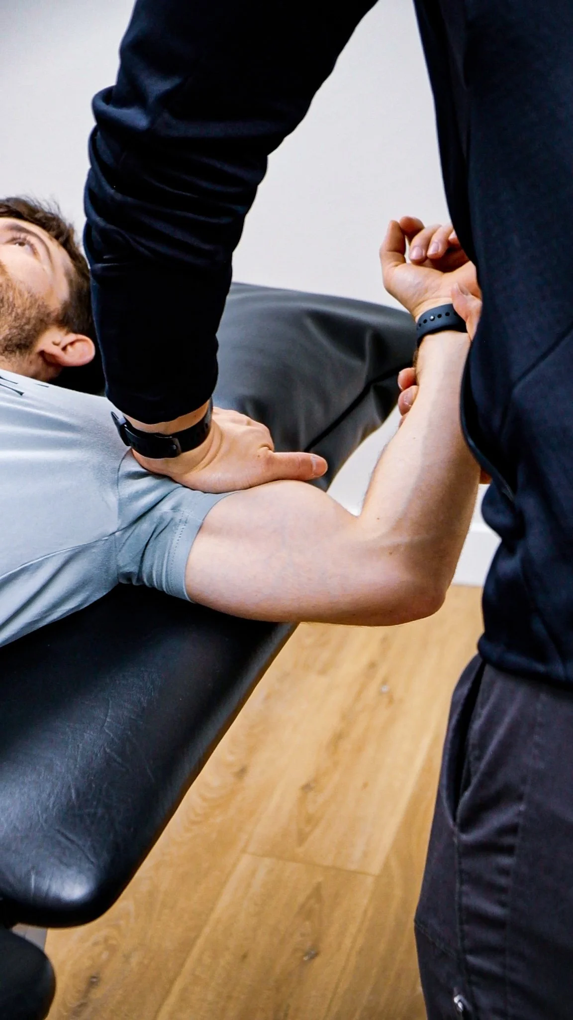 A person lying on a medical examination table with their arm raised and being examined or treated by another person standing next to them. The scene is in a physical therapy setting.