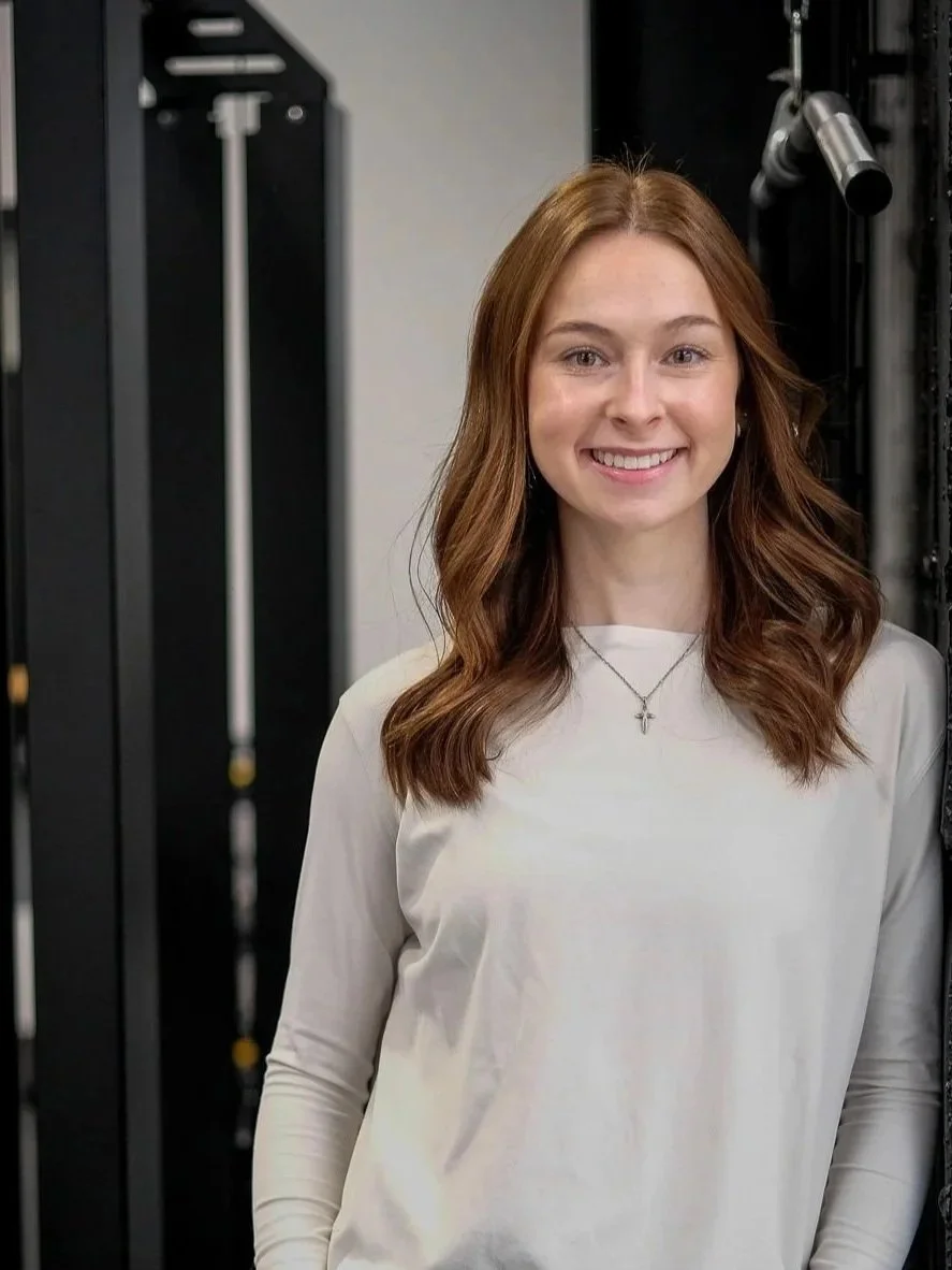 A young woman with long, wavy red hair smiling at the camera, standing in front of fitness equipment in a gym.