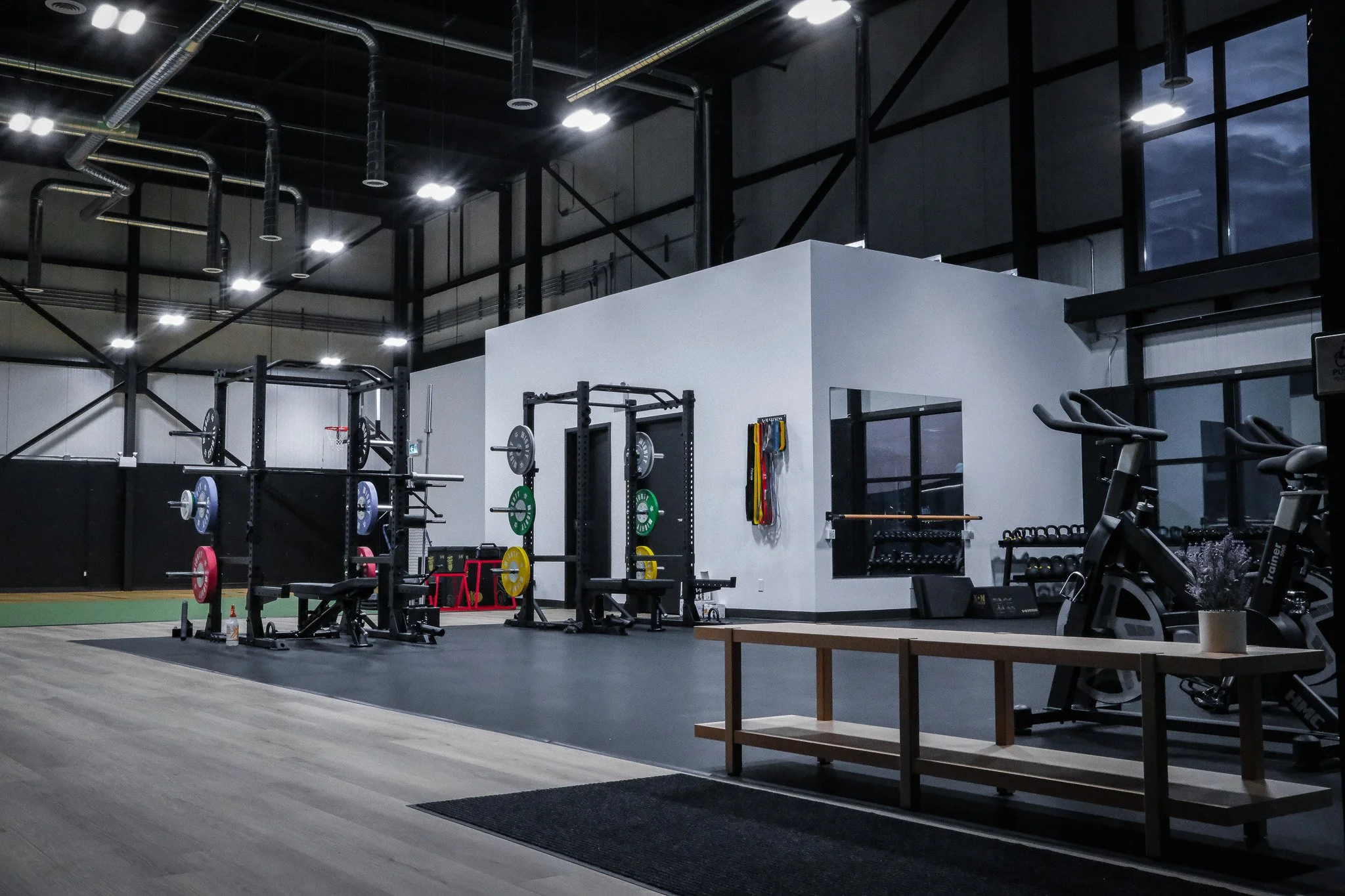 Empty gym with weightlifting equipment, including barbells with weight plates, squat racks, and cardio machines, inside a modern fitness center with high ceilings and large windows.