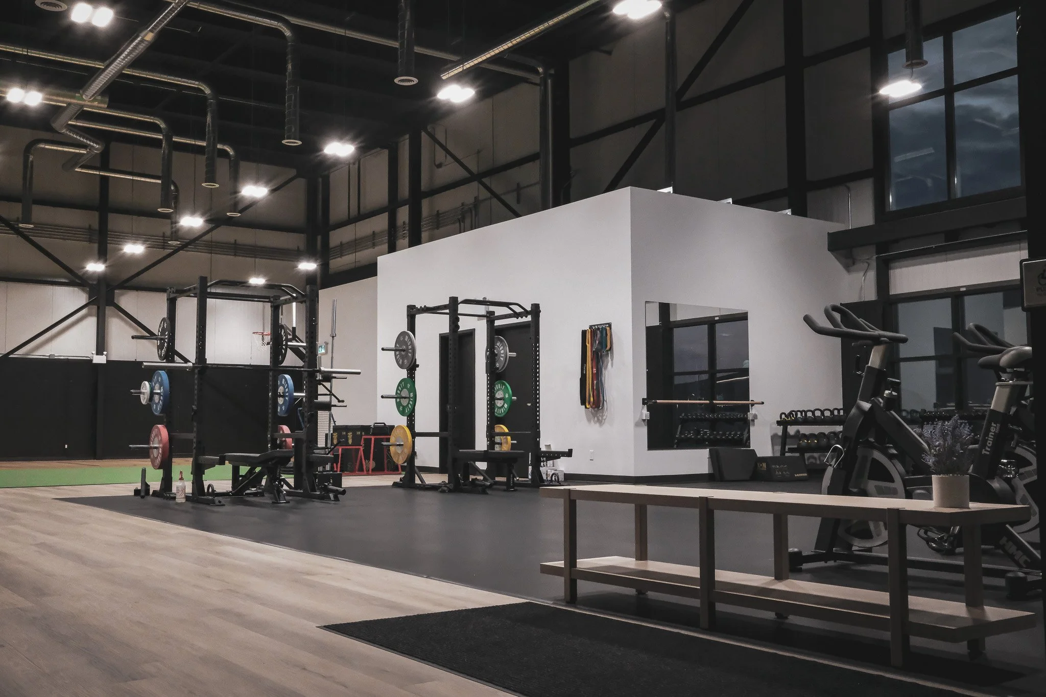 Interior of a modern gym space in a physiotherapy clinic with various workout equipment, including weightlifting stations, cardio machines, and mirrors. The gym has a high ceiling with visible industrial piping and large windows showing cloudy sky.