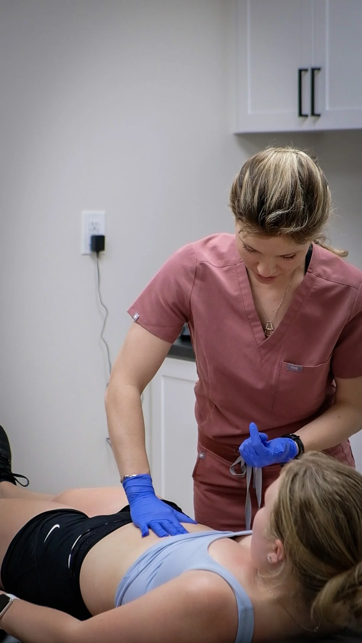 A pelvic health physiotherapist in pink scrubs and blue gloves performs a diastasis rectus abdominis assessment on a patient lying face up on a medical bed.
