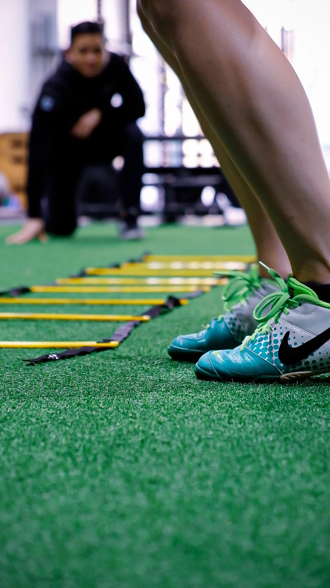 Close-up of a person's athletic shoes on green turf, with exercise agility ladder on the ground, in a fitness gym, with a sport physiotherapist crouching in the background.