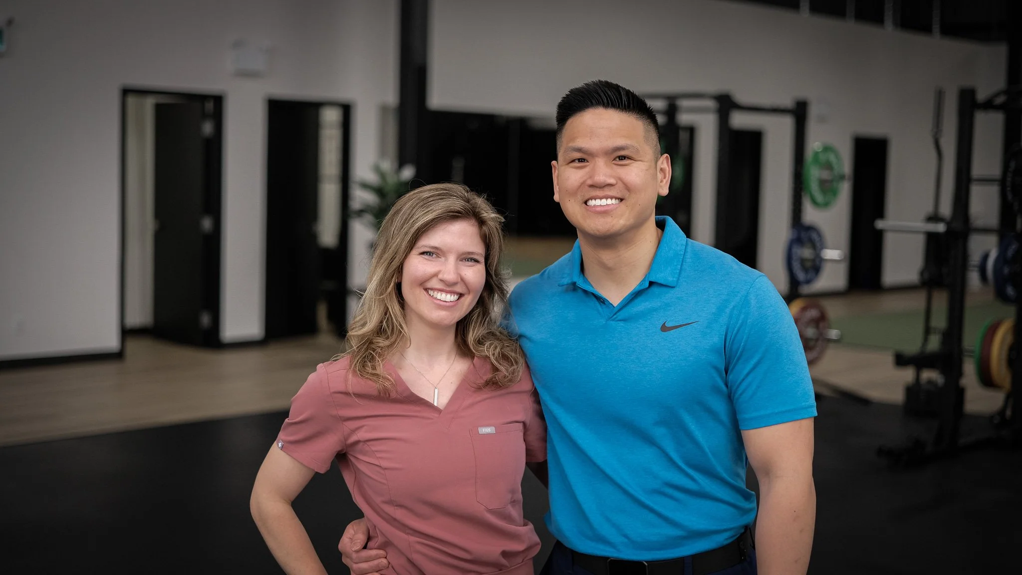 Two smiling people, a woman with wavy blonde hair wearing a pink scrubs and a man with short black hair wearing a blue polo shirt, standing in a gym with workout equipment in the background.
