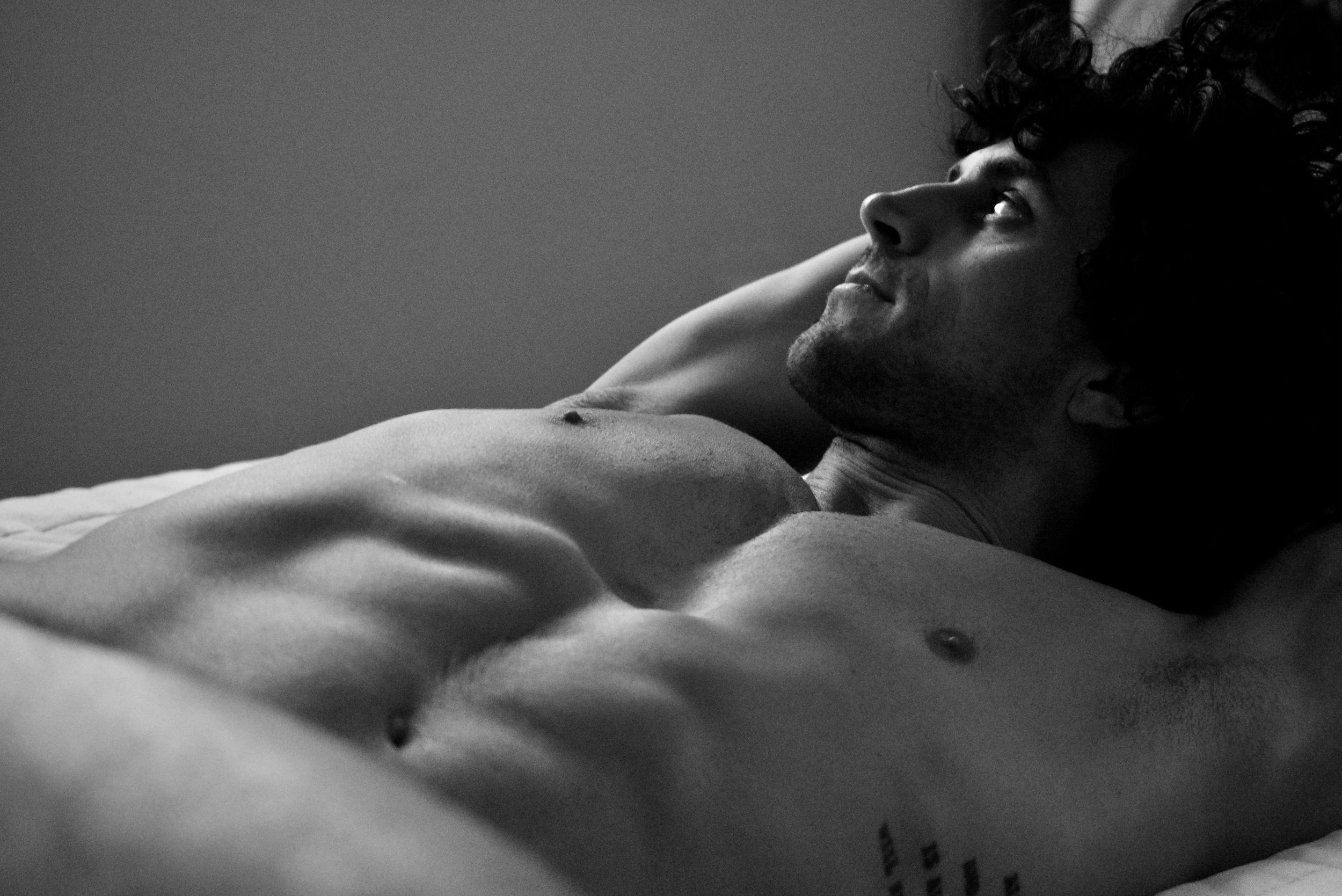 Black and white photograph of a smiling man with curly hair lying down and looking up, shirtless with a relaxed posture.