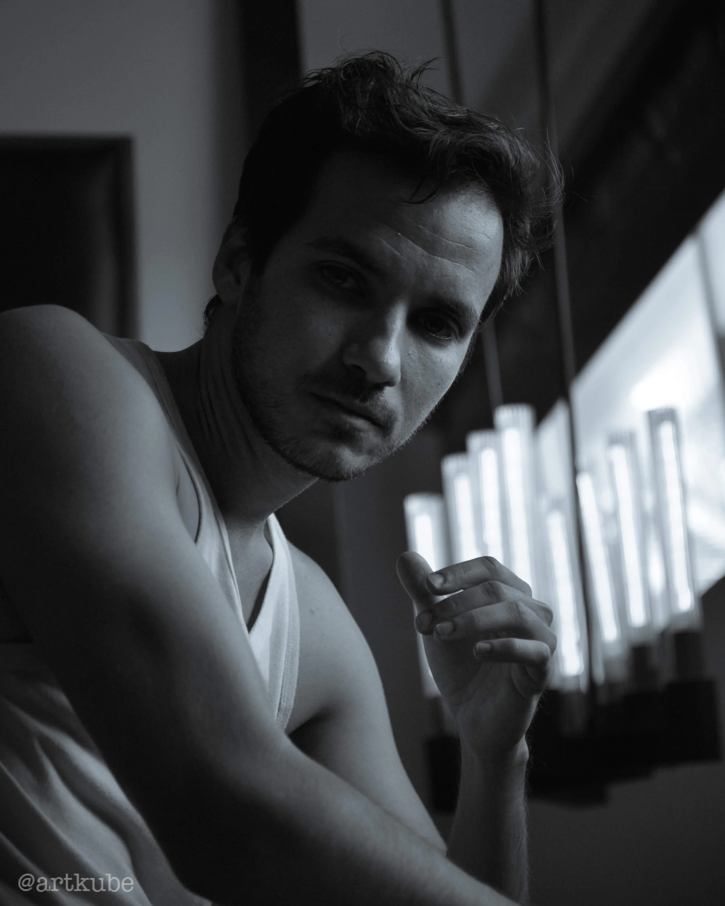 Black and white photo of a young man with dark, wavy hair, wearing a sleeveless shirt, looking at the camera while holding a small object in his right hand, with a window in the background.