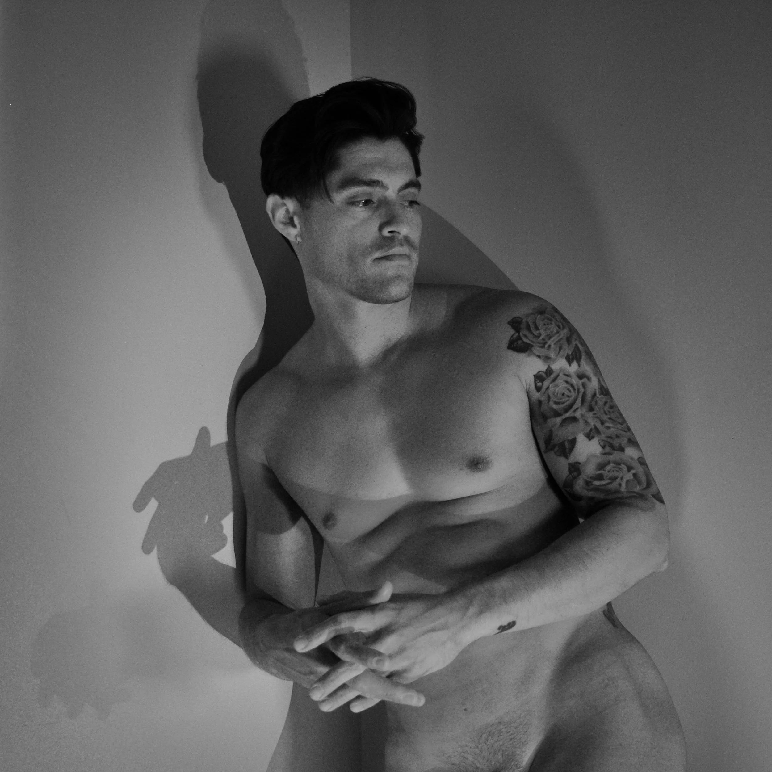 Black and white photo of a man with tattoos on his arm, standing against a wall, looking to the side.