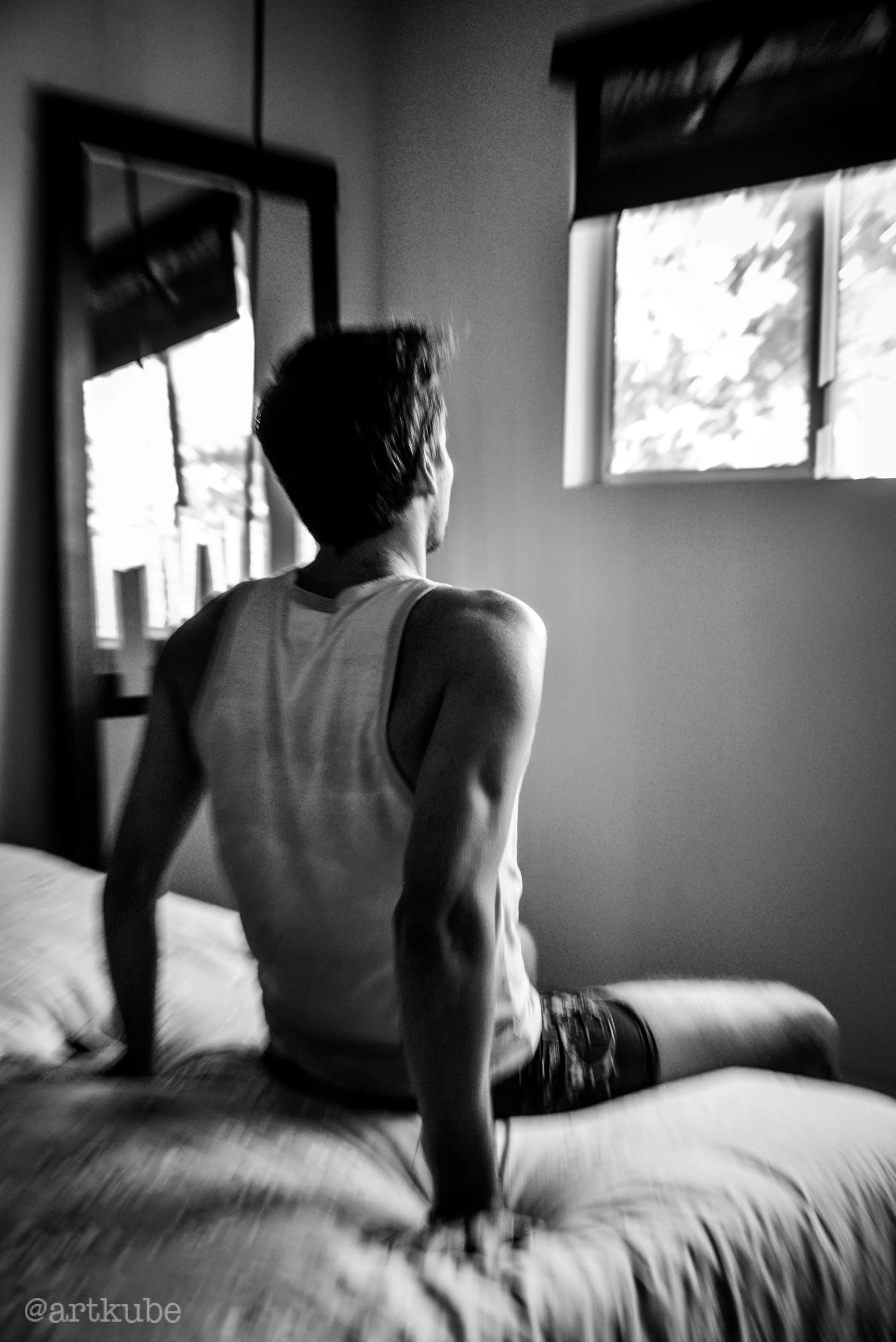 A person sitting on a bed in a room with a window, seen from behind. The person is wearing a tank top and patterned shorts, and the room has a mirror on the wall with a reflection. The photo is in black and white with a cinematic, artistic style.
