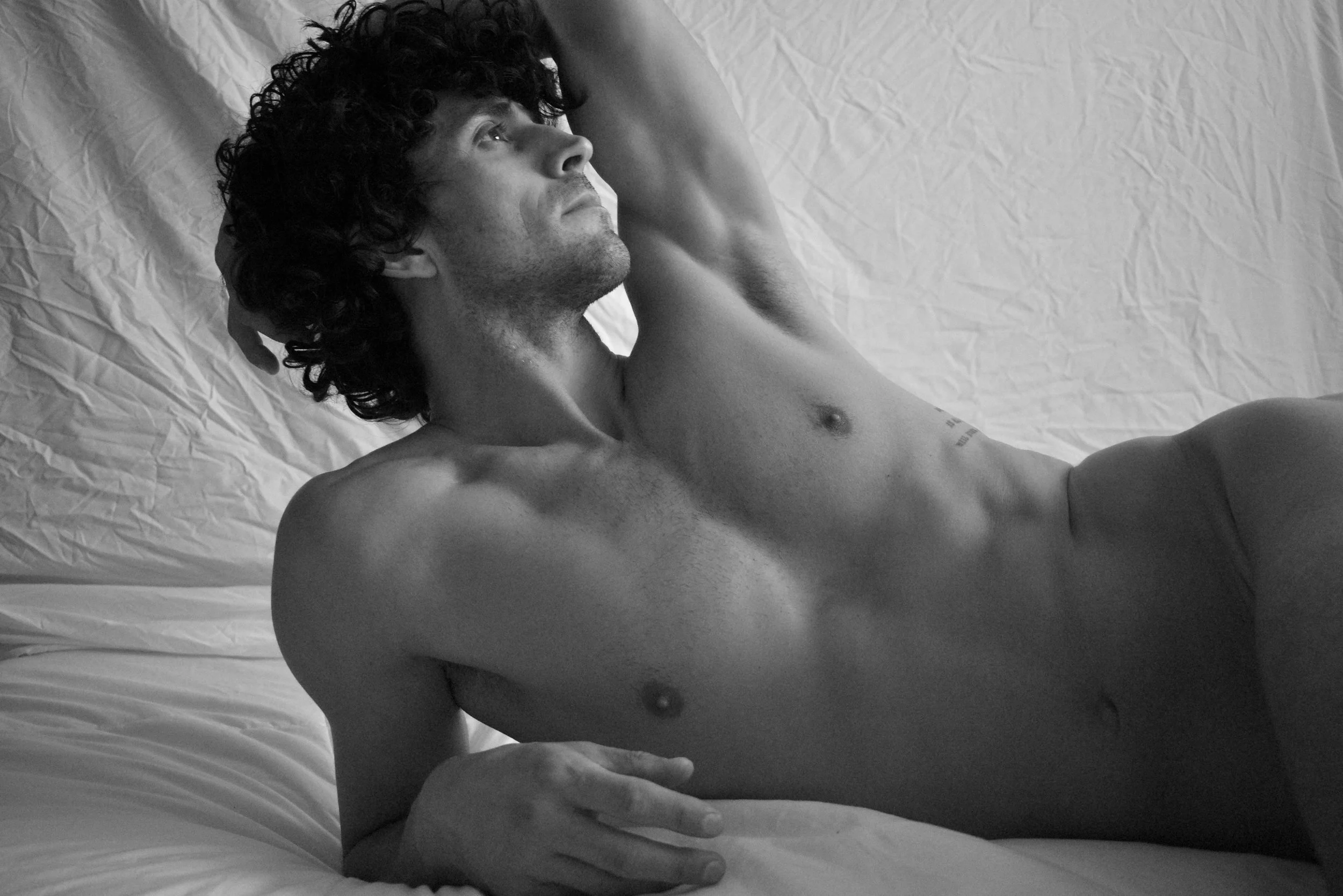 A shirtless man with curly hair lying on a bed, looking to the side with his left hand resting on his head.