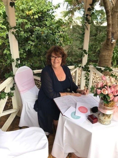 Wedding celebrant seated at a ceremony signing table with flowers and paperwork.