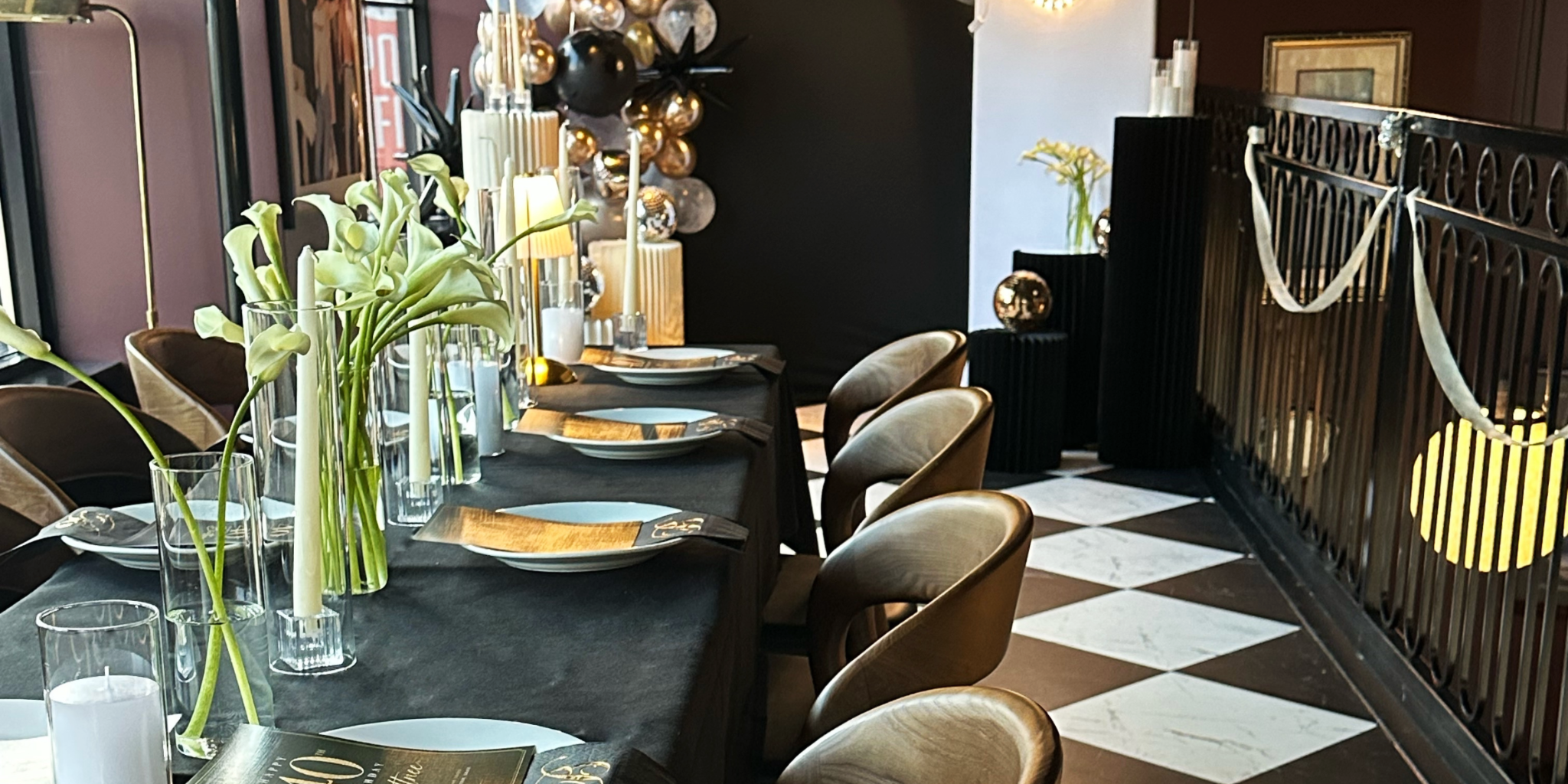 Elegant dining table set with white calla lilies in glass vases, surrounded by beige chairs, with candles and decorative balls on a black wall, and a black and white checkered floor in a stylish restaurant interior.