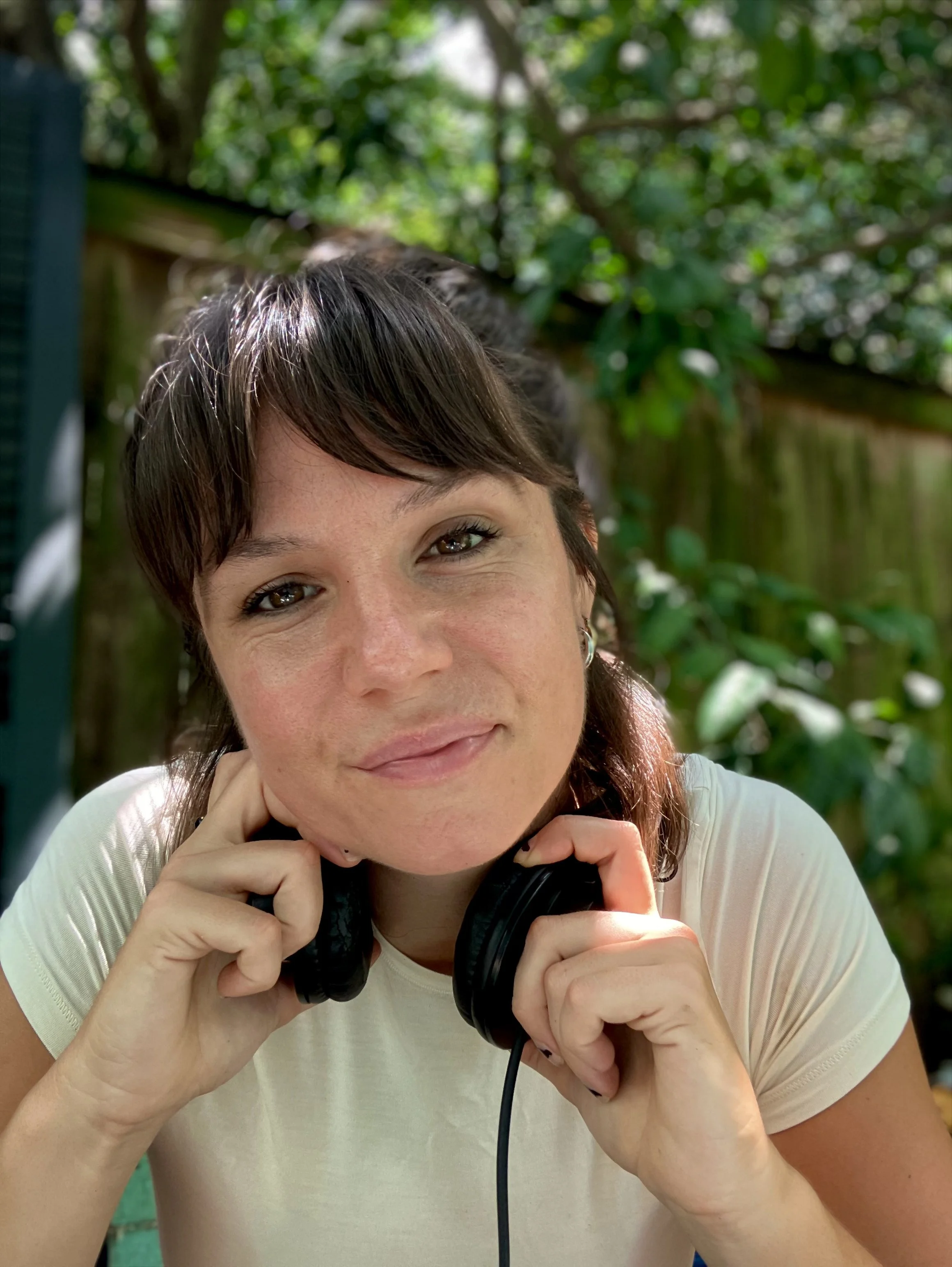 A woman with short dark hair, brown eyes, and earrings, smiling outdoors with green foliage in the background. She is holding black headphones near her neck with both hands.