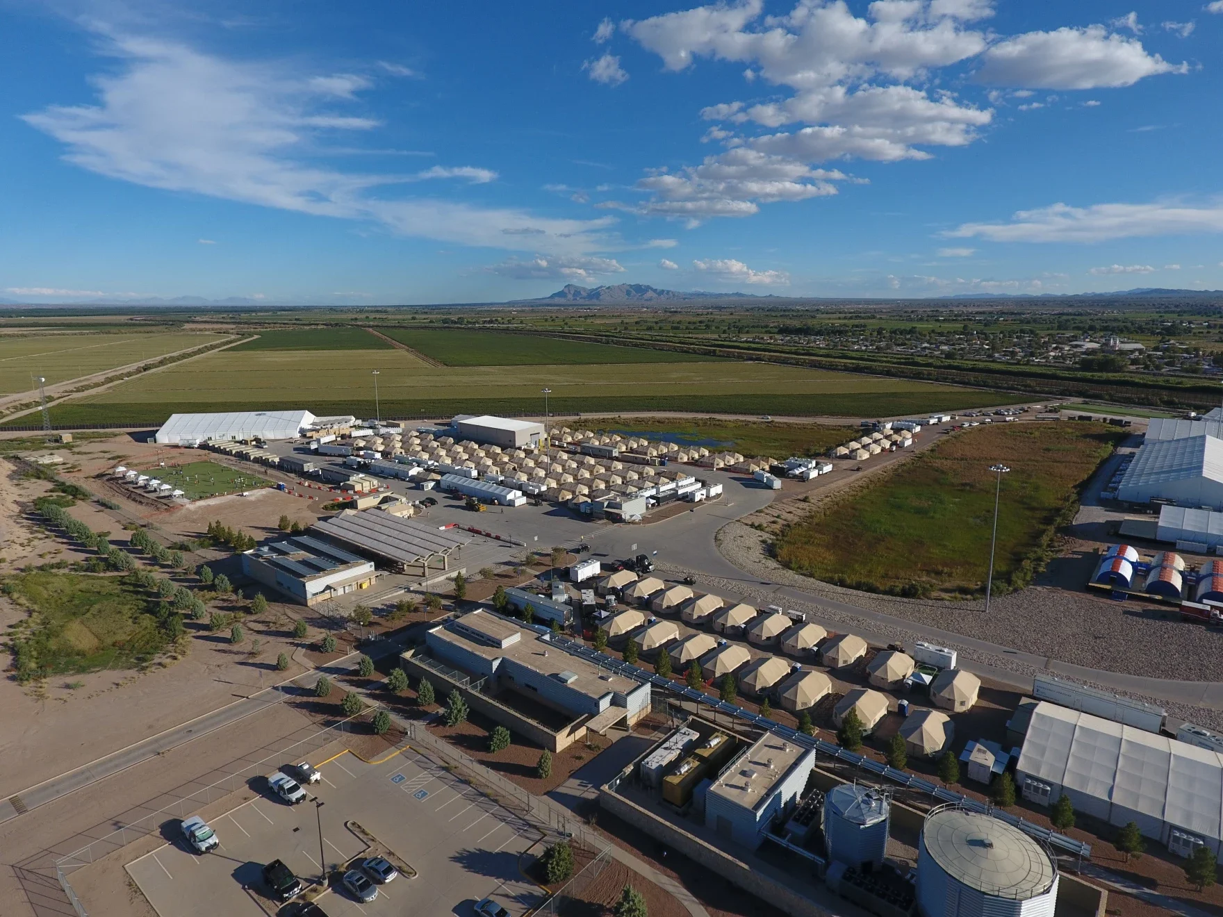 Aerial view of a large event setup with tents and temporary structures in a rural area, surrounded by fields and distant mountains under a partly cloudy sky.