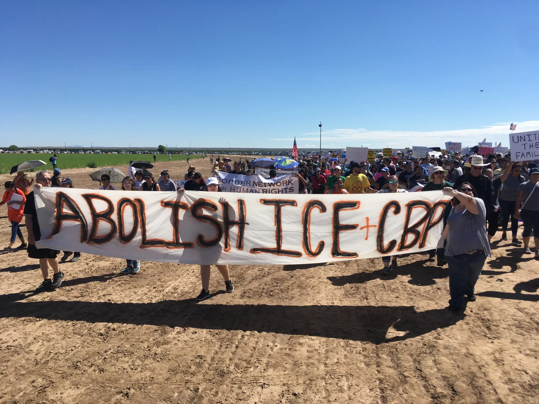 A large group of protesters marching outdoors on a sunny day, holding signs and banners, including one prominent banner that reads 'ABOLISH ICE + CBP'. Some protesters have umbrellas to shield themselves from the sun. The setting appears to be an open area with a grassy field and a parking lot in the background.