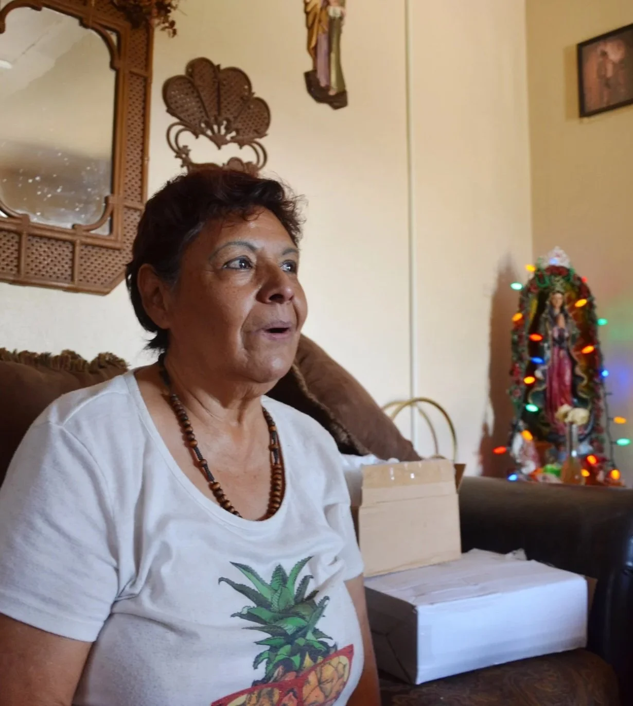A woman with short dark hair and a beaded necklace sits on a couch in a living room, wearing a white t-shirt with a pineapple design. Behind her, a wooden mirror, and a Virgen de Guadalupe statue with colorful Christmas lights.