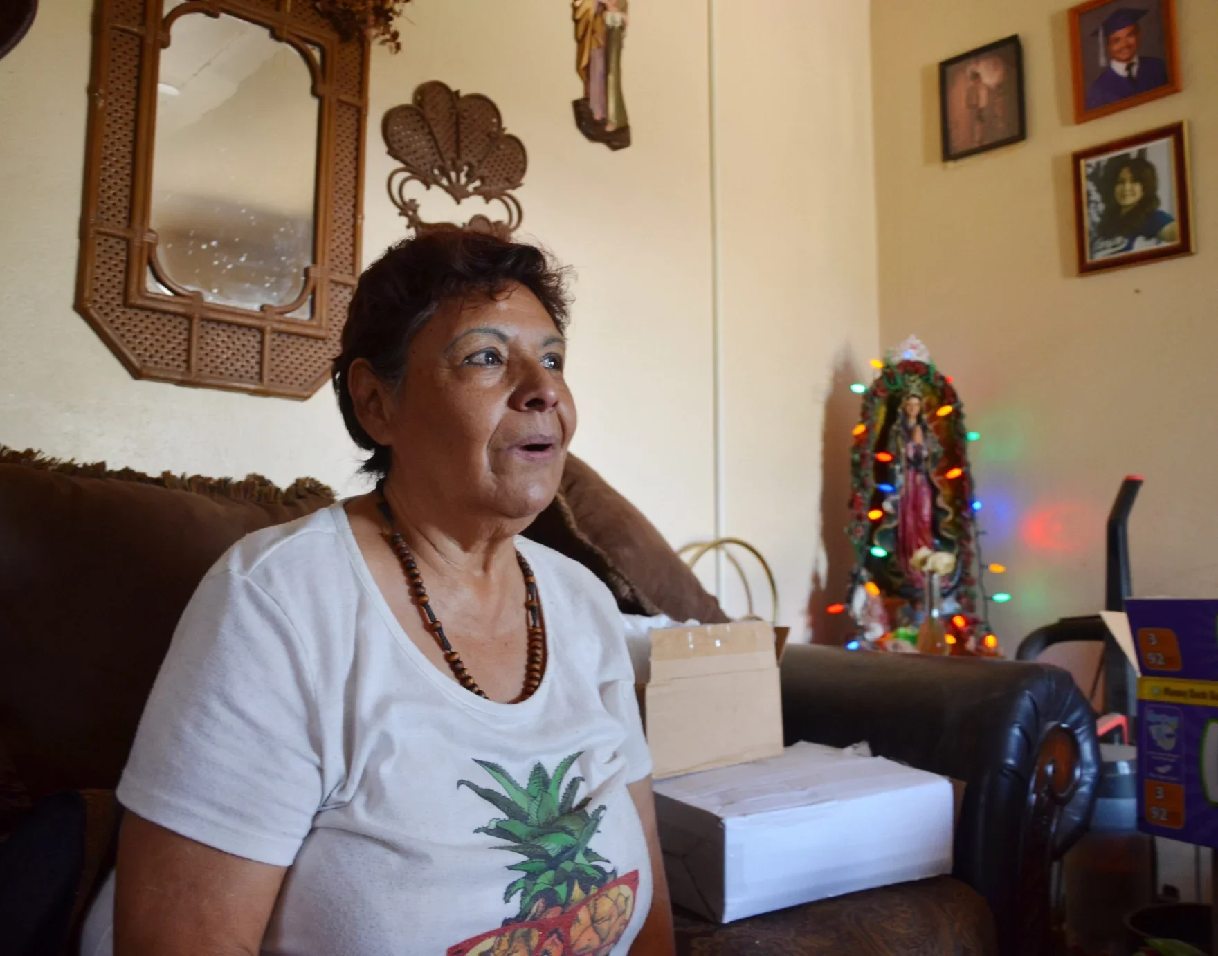 A woman with short dark hair and a beaded necklace sits on a couch in a living room, wearing a white t-shirt with a pineapple design. Behind her, a wooden mirror, and a Virgen de Guadalupe statue with colorful Christmas lights.