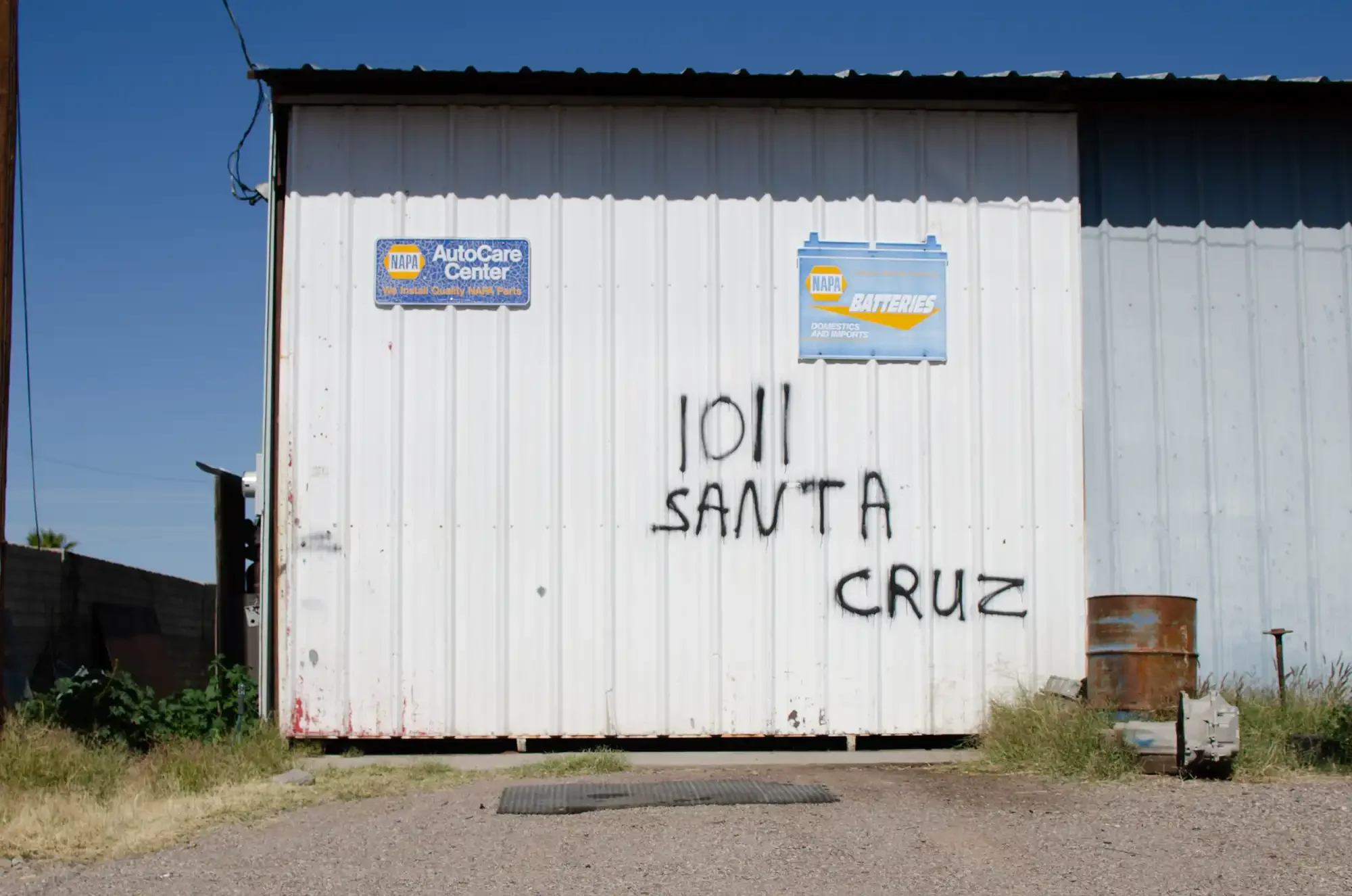The image shows a white metal building displaying signs for an auto care business in Presidio, Texas. There is graffiti on the building that reads '1011 SANTA CRUZ'.