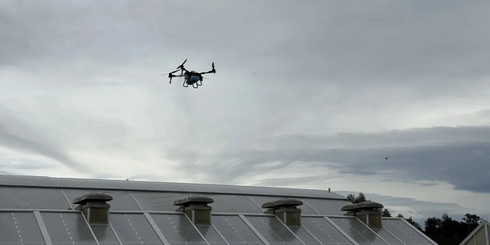 A drone flying above a building with solar panels and ventilation vents on the roof against a cloudy sky.
