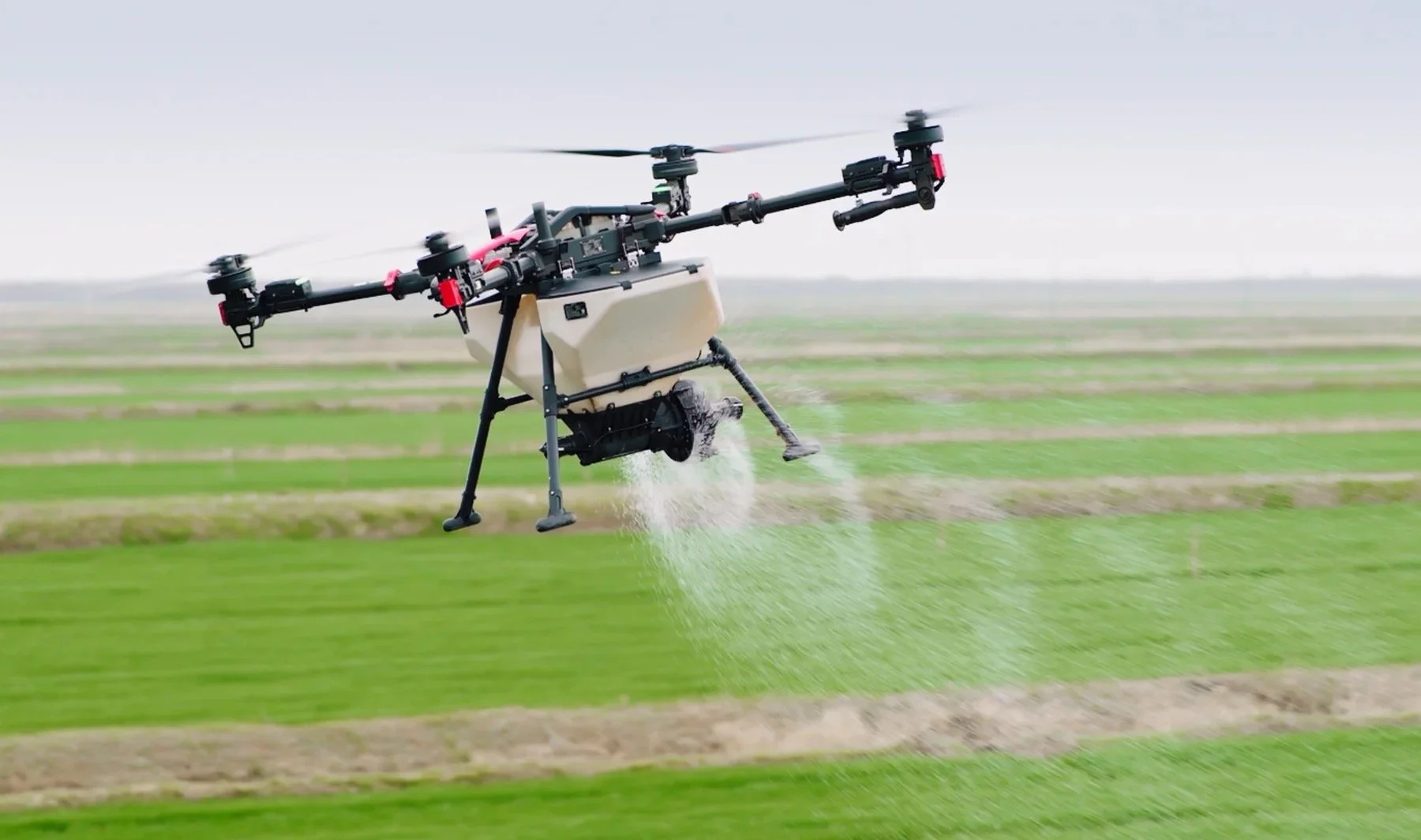 A large agricultural drone flying over green fields, dispersing liquid or fertilizer through a spray system.