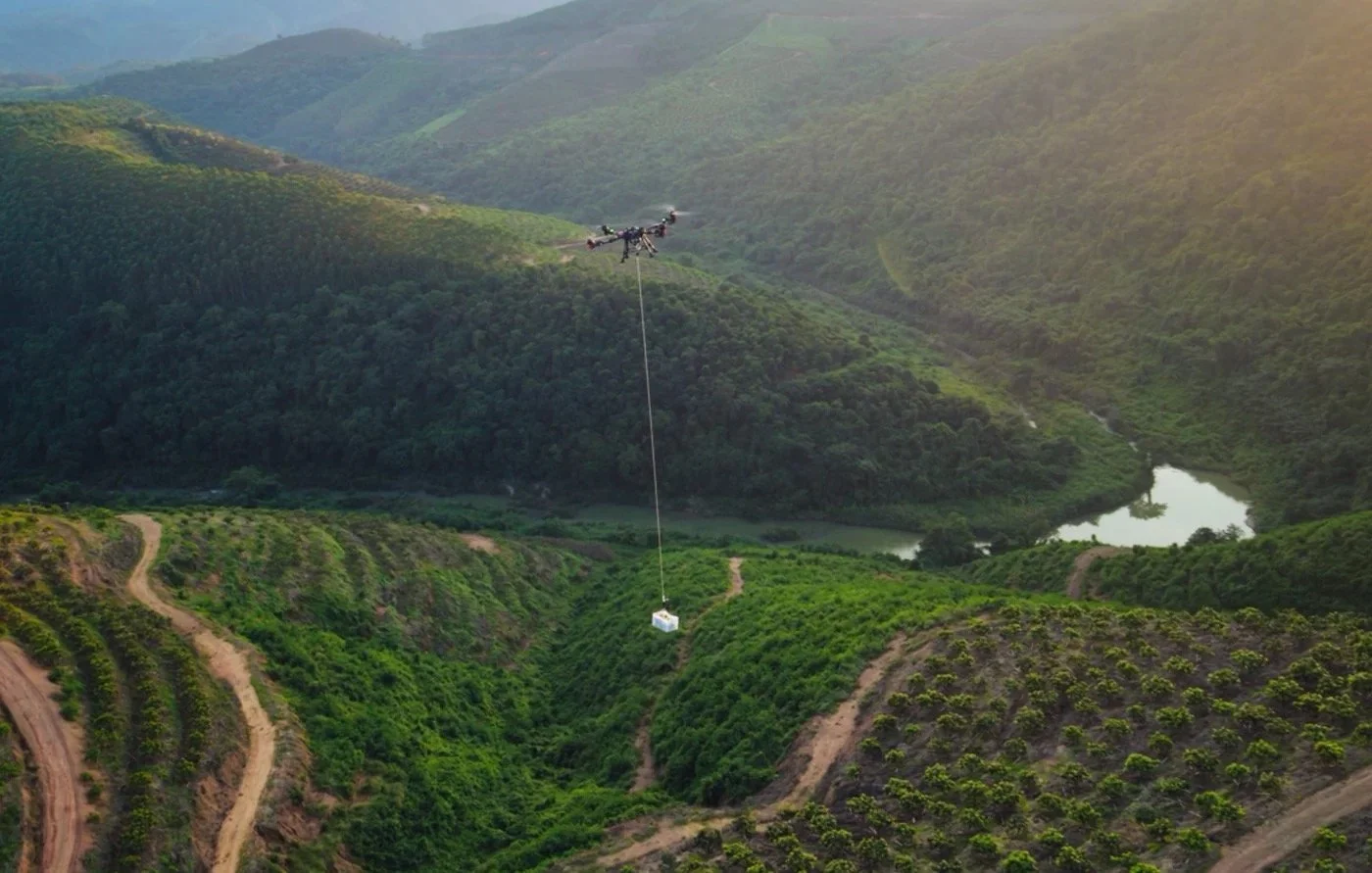 A drone carrying a package flying over a green hilly landscape with a river in the background.