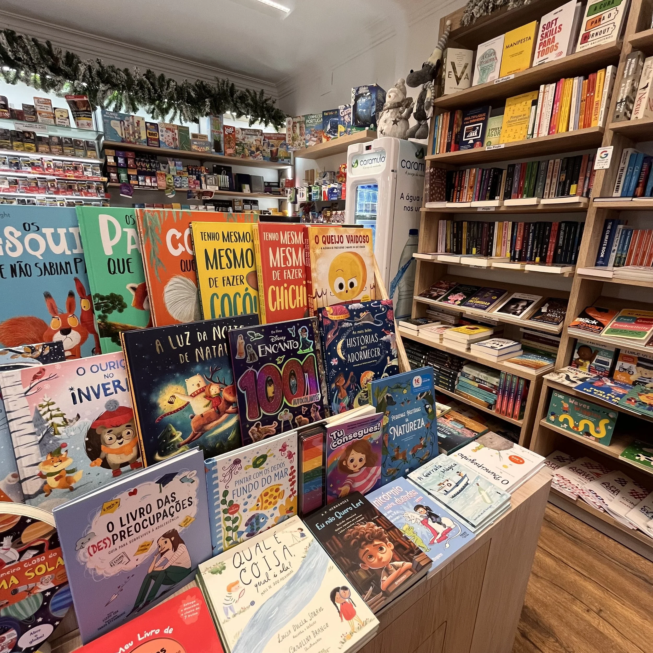 Imagem de uma livraria com estantes cheias de livros e livros infantis na mostra, com títulos em português e uma decoração de Natal com guirlanda na parte superior da prateleira.