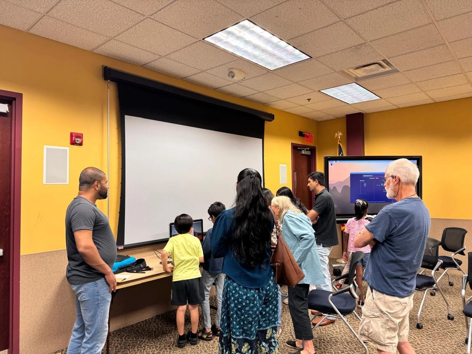 Group of children and adults gathered in a classroom or meeting room, standing near a large blank projection screen and a monitor displaying a game of Monopoly.