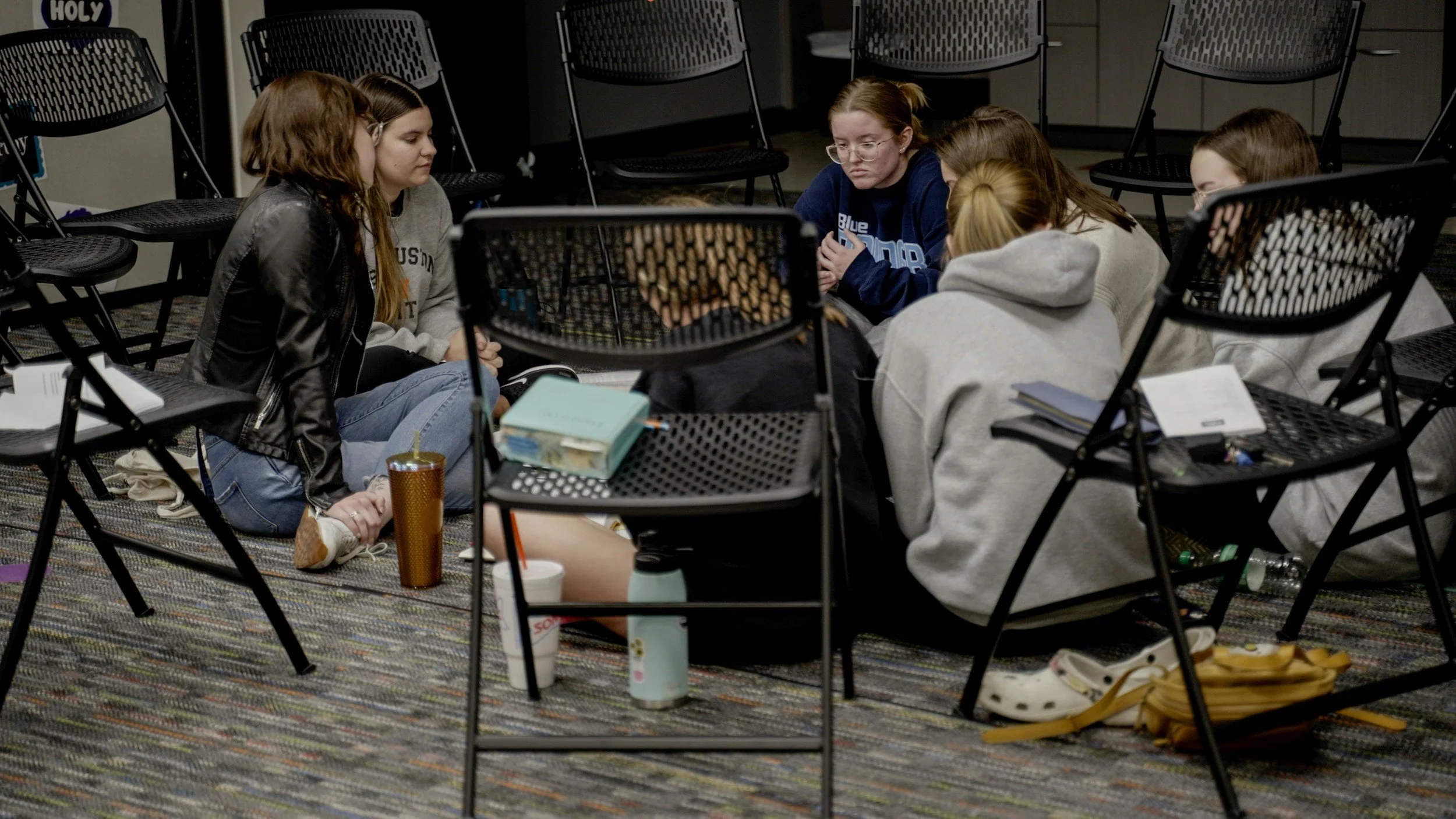 Group of young women sitting cross-legged on the floor in a circle, engaged in a discussion, with chairs arranged around them and personal items like books, drinks, and backpacks nearby.