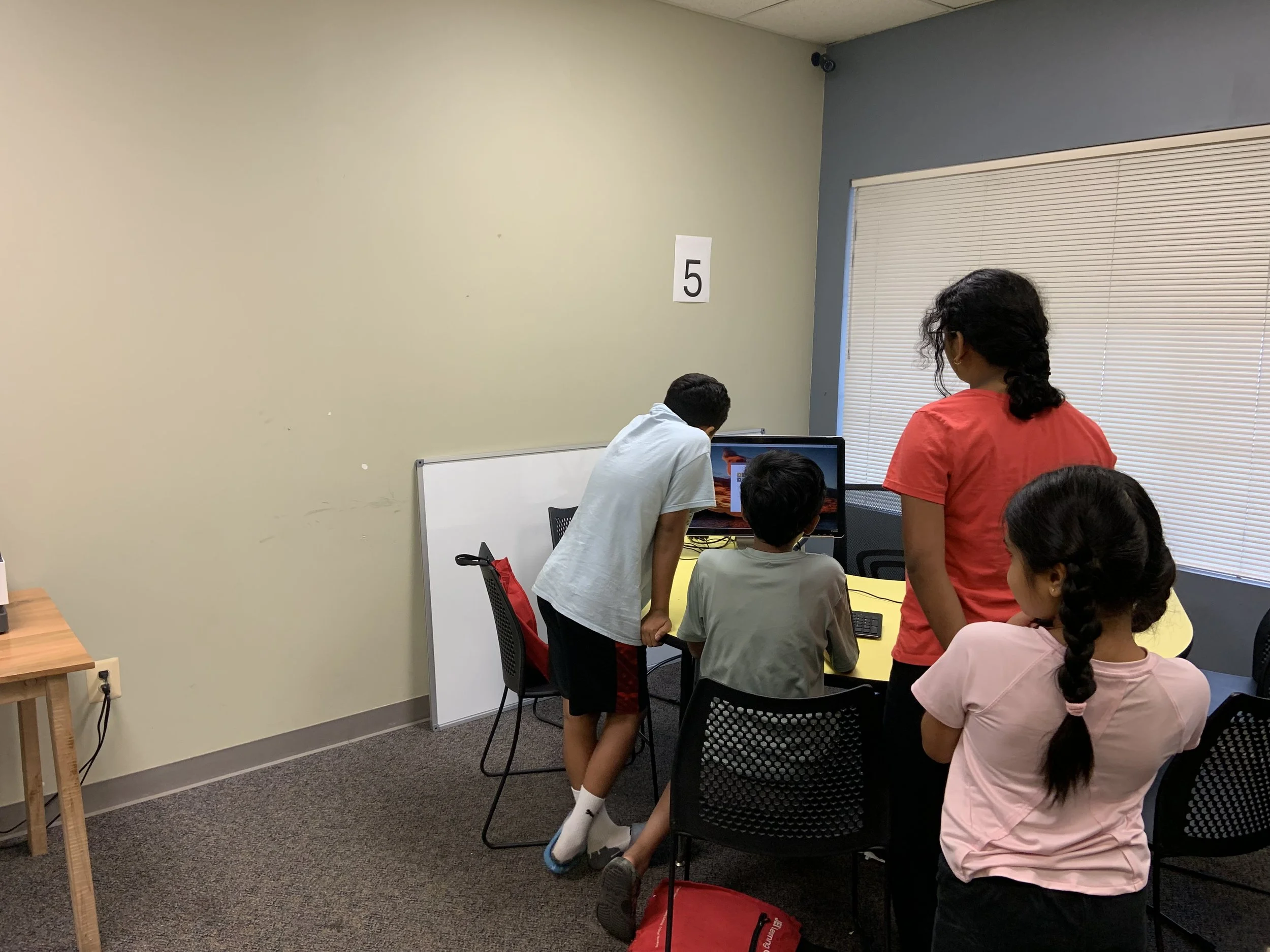 Children gathered around a computer in a classroom, with one girl standing and the others seated. The classroom has a whiteboard, beige walls, and a window with blinds.