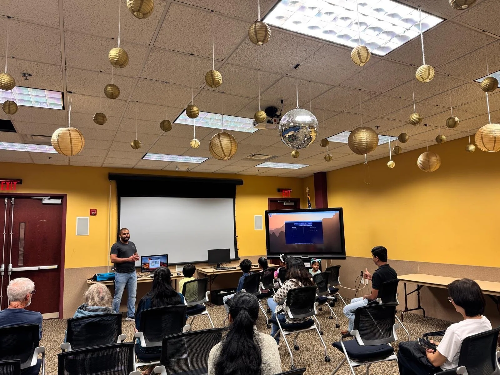 A classroom or conference room where a man is giving a presentation to a diverse group of people. The room has yellow walls, a large projection screen, a digital monitor, and decorative hanging paper lanterns and a disco ball on the ceiling.
