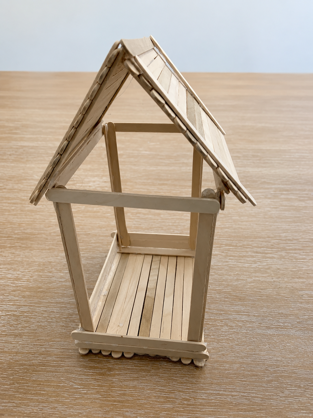 Small wooden dollhouse with a gabled roof, open front, wooden floor, and porch, placed on a wooden surface.