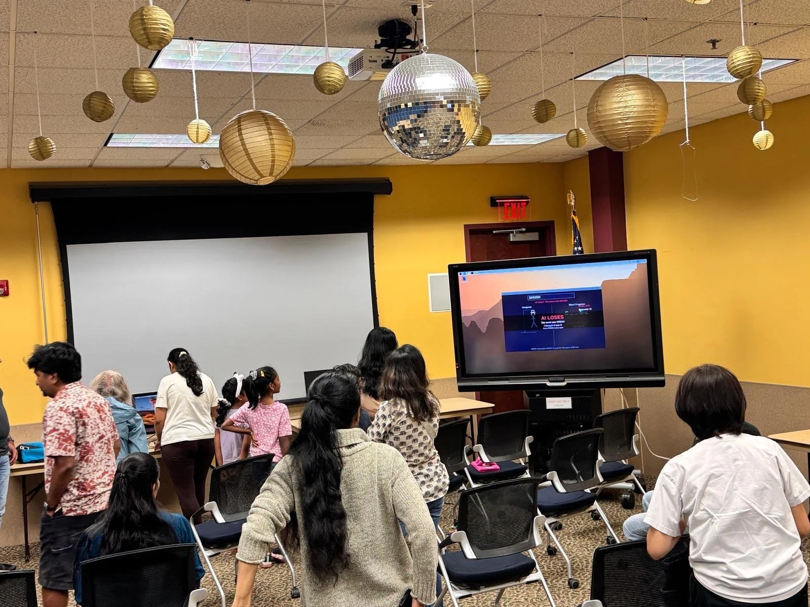 A room filled with people, including children and adults, gathering around chairs and tables. There is a large projection screen at the front and a display monitor showing a game on a computer. The ceiling has hanging gold paper lanterns and a disco 