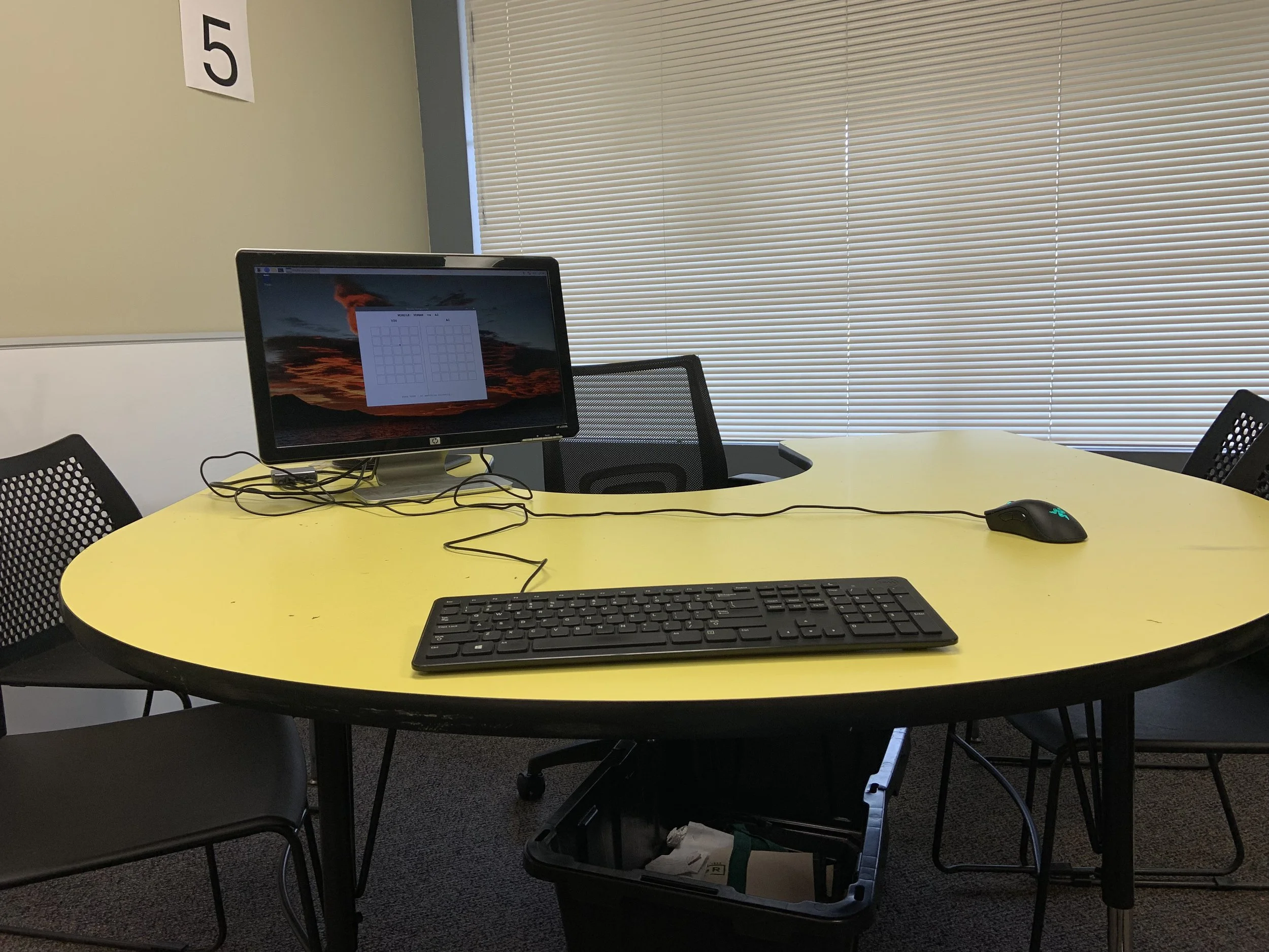 An office conference room with a yellow oval table, black mesh chairs, a computer monitor, a keyboard, and a mouse on the table. There is a wall with blinds covering a window and a sign with the number 5 on the wall.