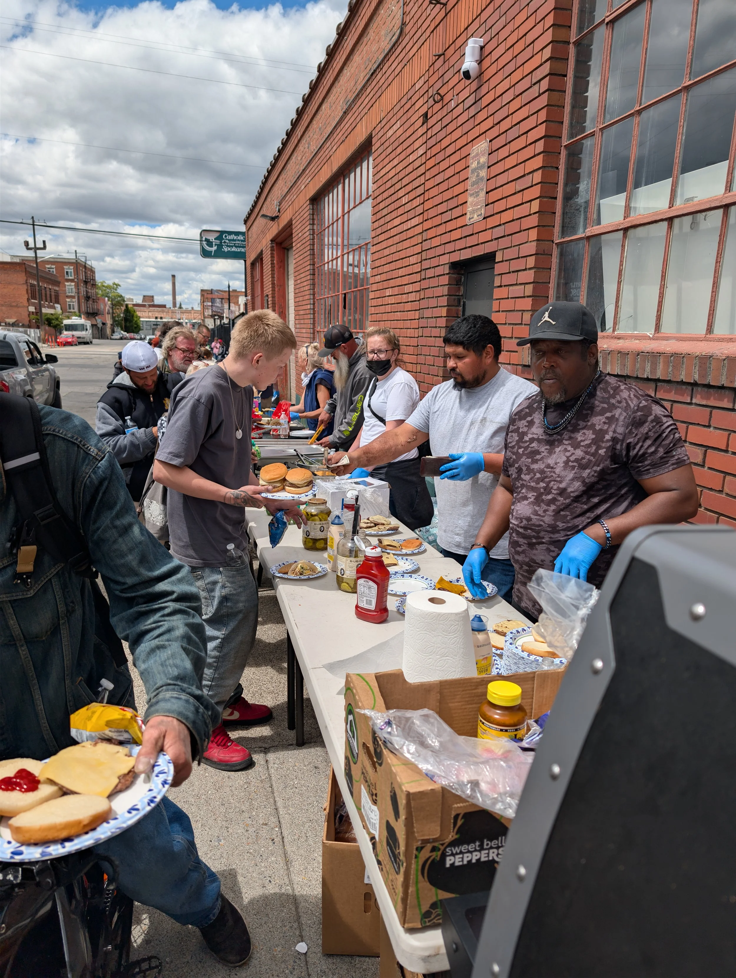 People lined up at an outdoor food distribution event on a sidewalk beside a brick building, with tables of food and supplies including hamburgers,, condiments, and paper towels.