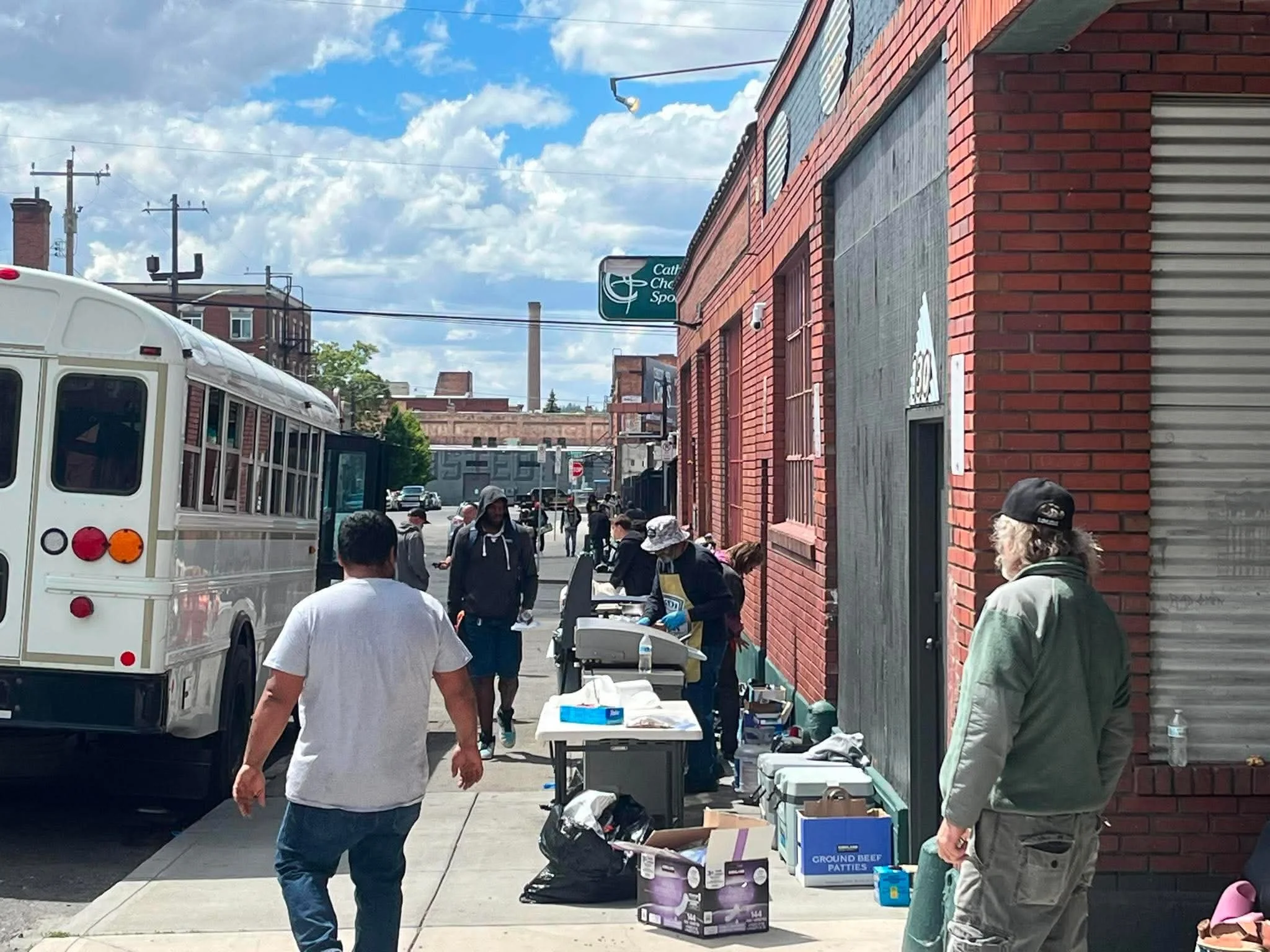 People walking along a street sidewalk with tables and boxes for an outdoor sale or event, next to red brick buildings under a partly cloudy sky.