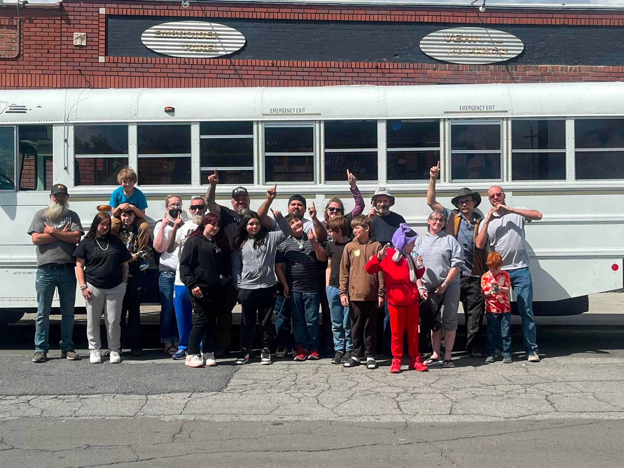 A group of people, including men, women, and children, standing in front of a white bus all of them volunteered to feed homeless, parked on a street Downtown Spokane. they are pointing up to God who made it possible.
