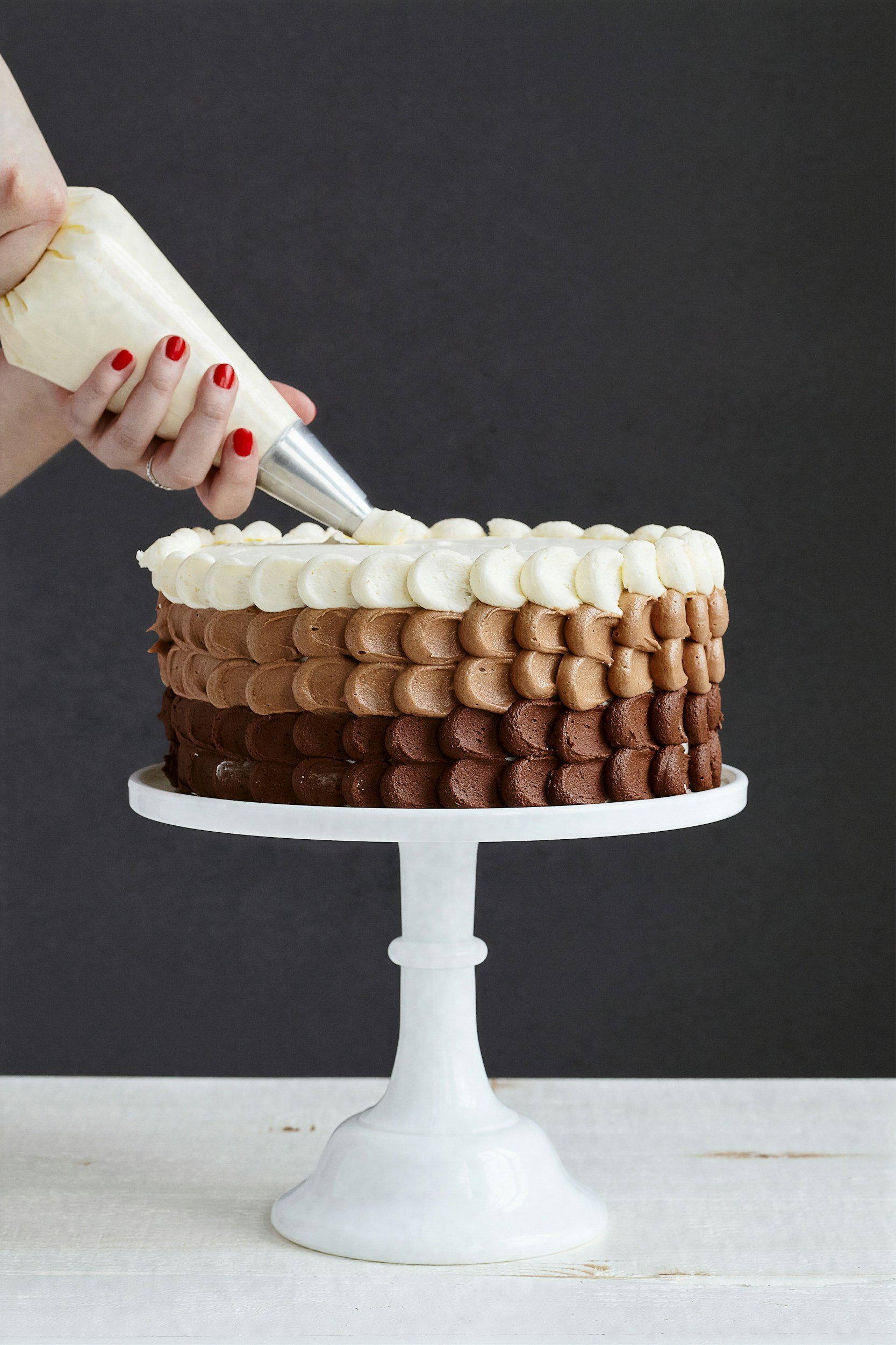 A multi-layered chocolate and vanilla cake on a white cake stand, with white and chocolate frosting, and a person piping white frosting decoration on top.