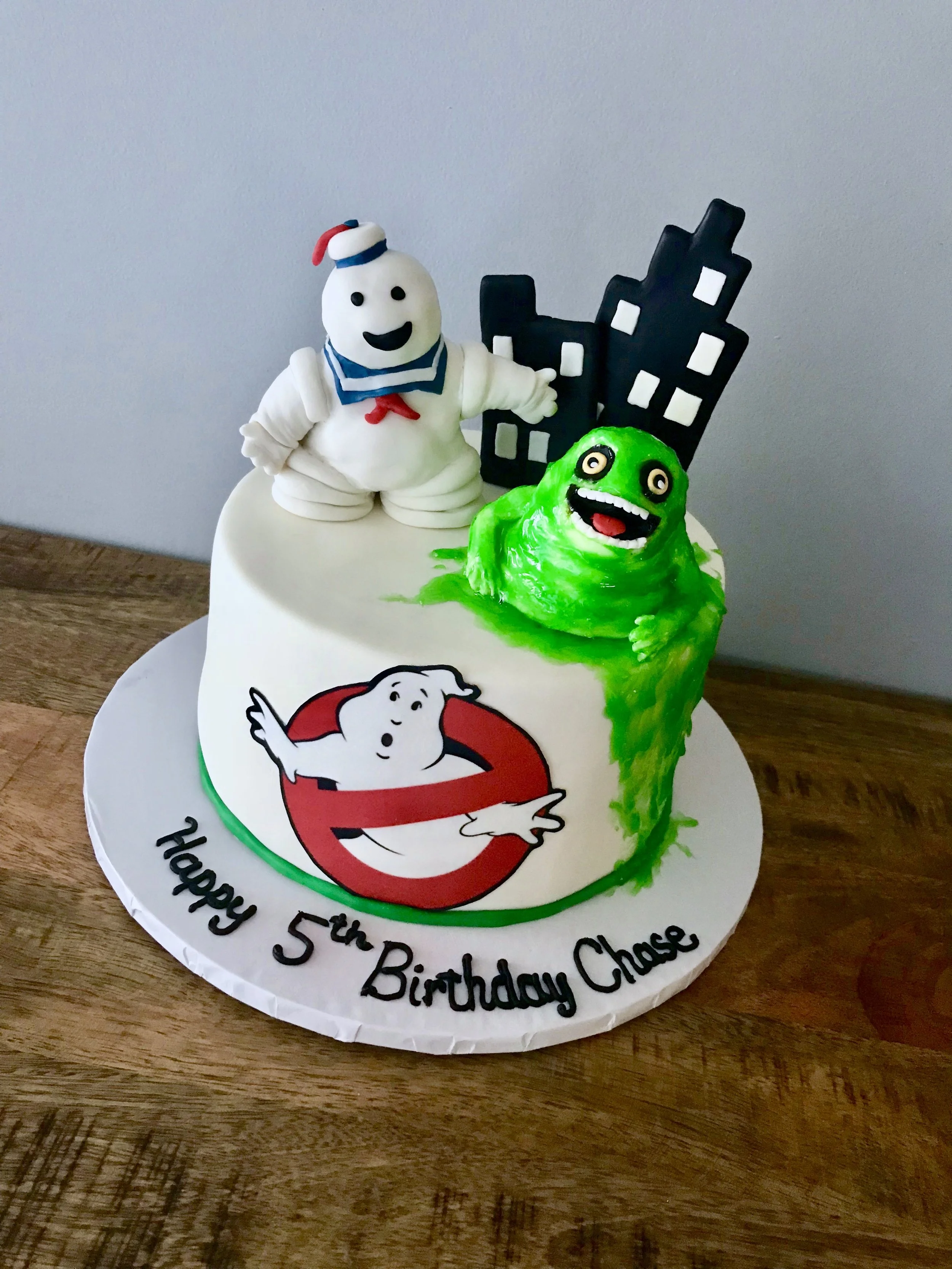 A birthday cake decorated with a ghost from Ghostbusters, a green slime monster, and a cityscape silhouette, with a message for a fifth birthday celebration.