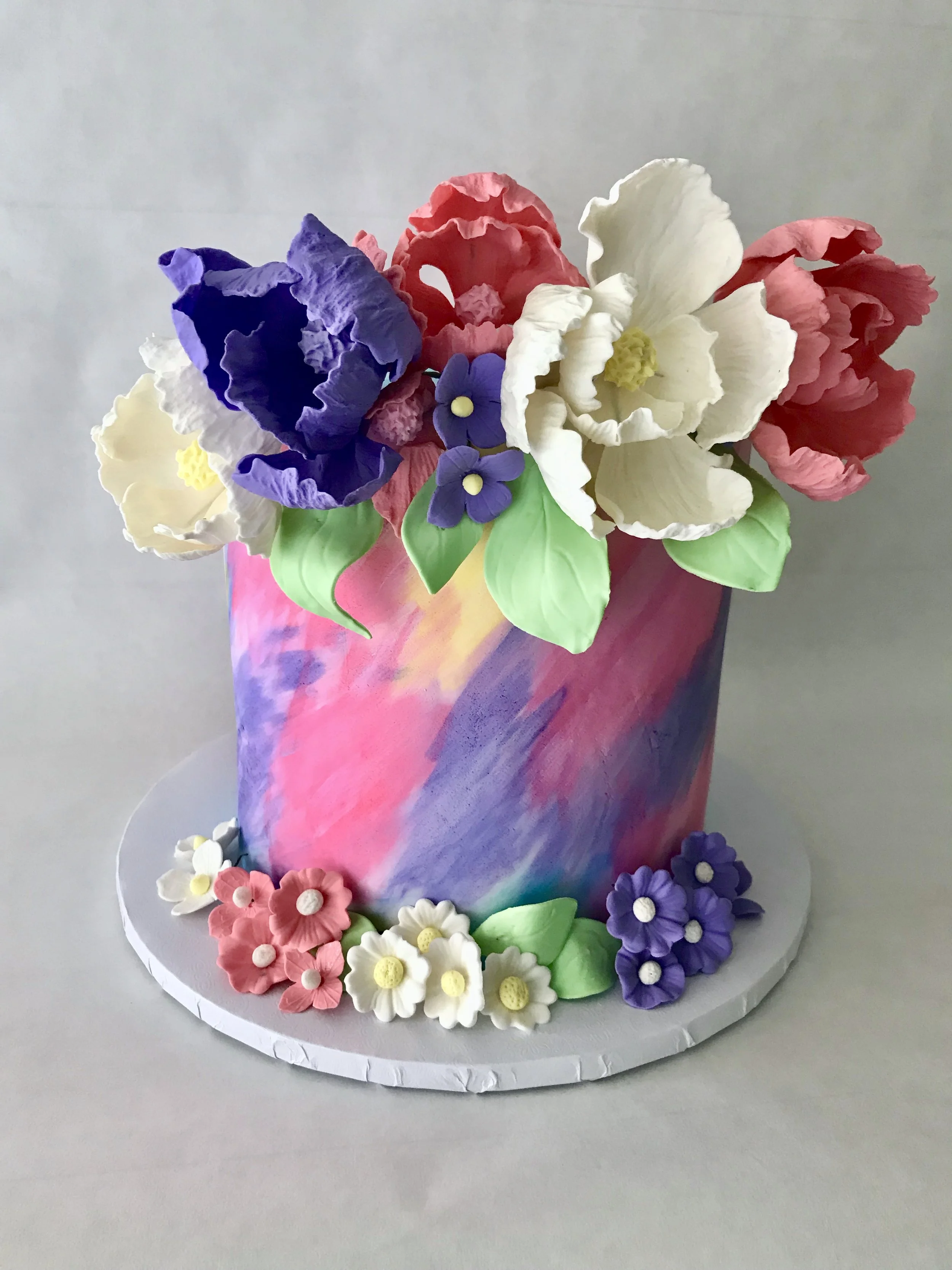 Colorful birthday cake with smooth rainbow-colored frosting topped with edible sugar flowers in purple, white, pink, red, and yellow, decorated with sugar leaves and flowers around the base, placed on a round white cake board.