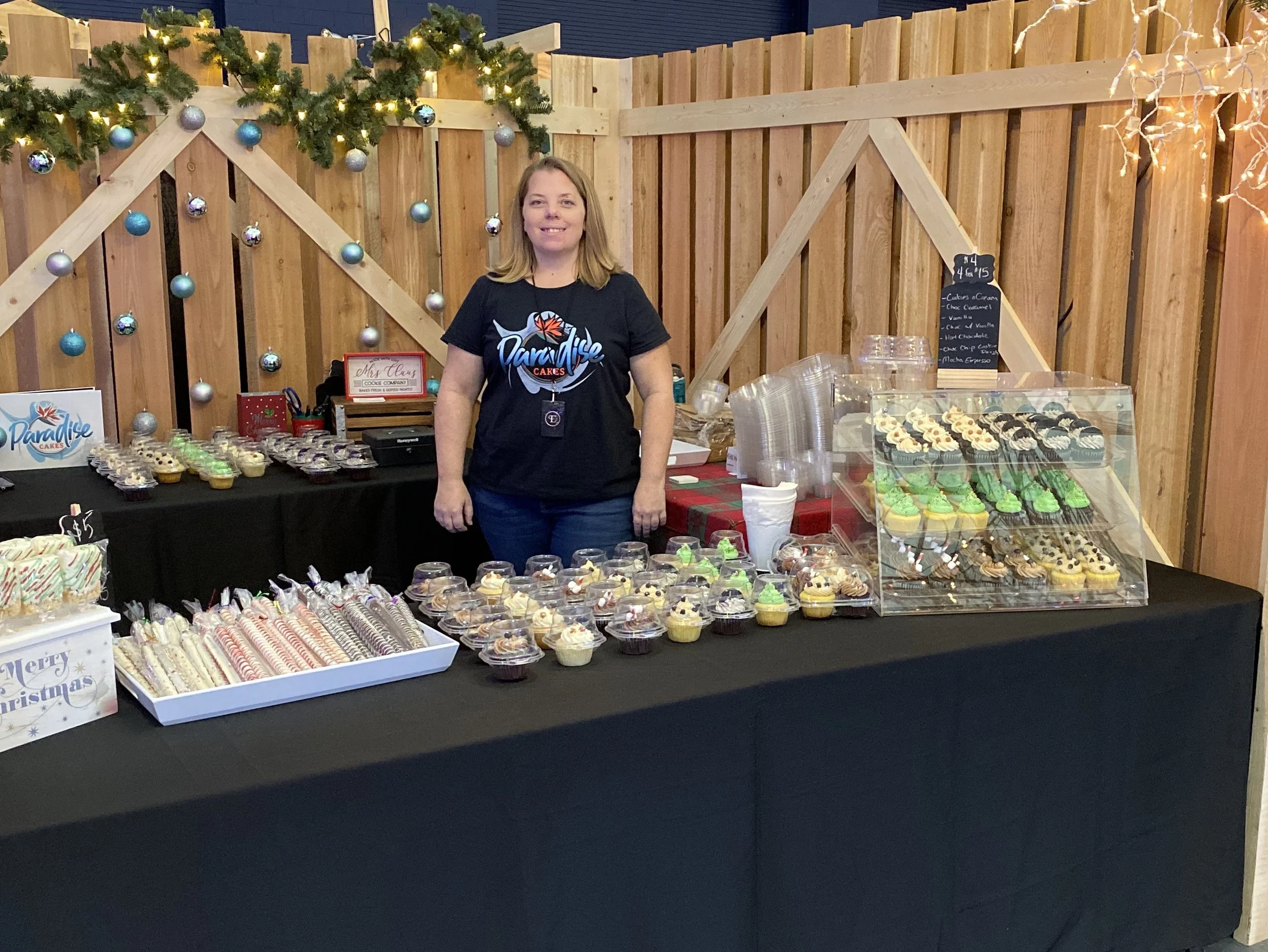 Smile of a woman standing behind a table of cupcakes decorated with icing, sprinkles, and fruit, at a holiday-themed booth with Christmas ornaments and garlands.