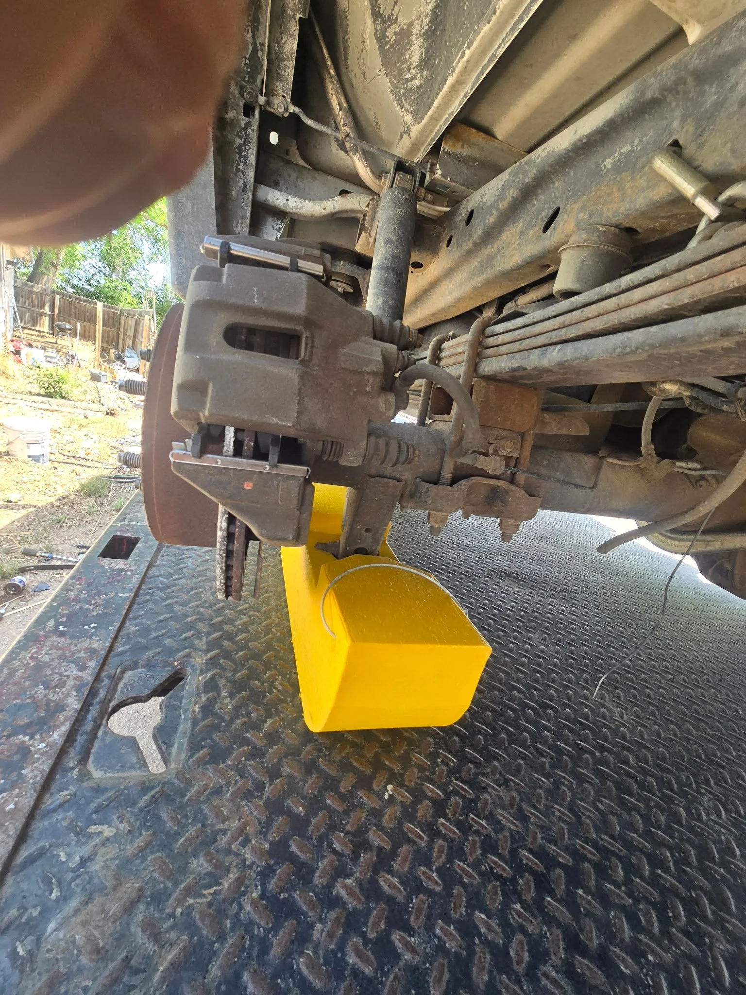 Underside of a vehicle with suspension components, brake caliper, and brake disc visible, with a yellow block supporting the vehicle.