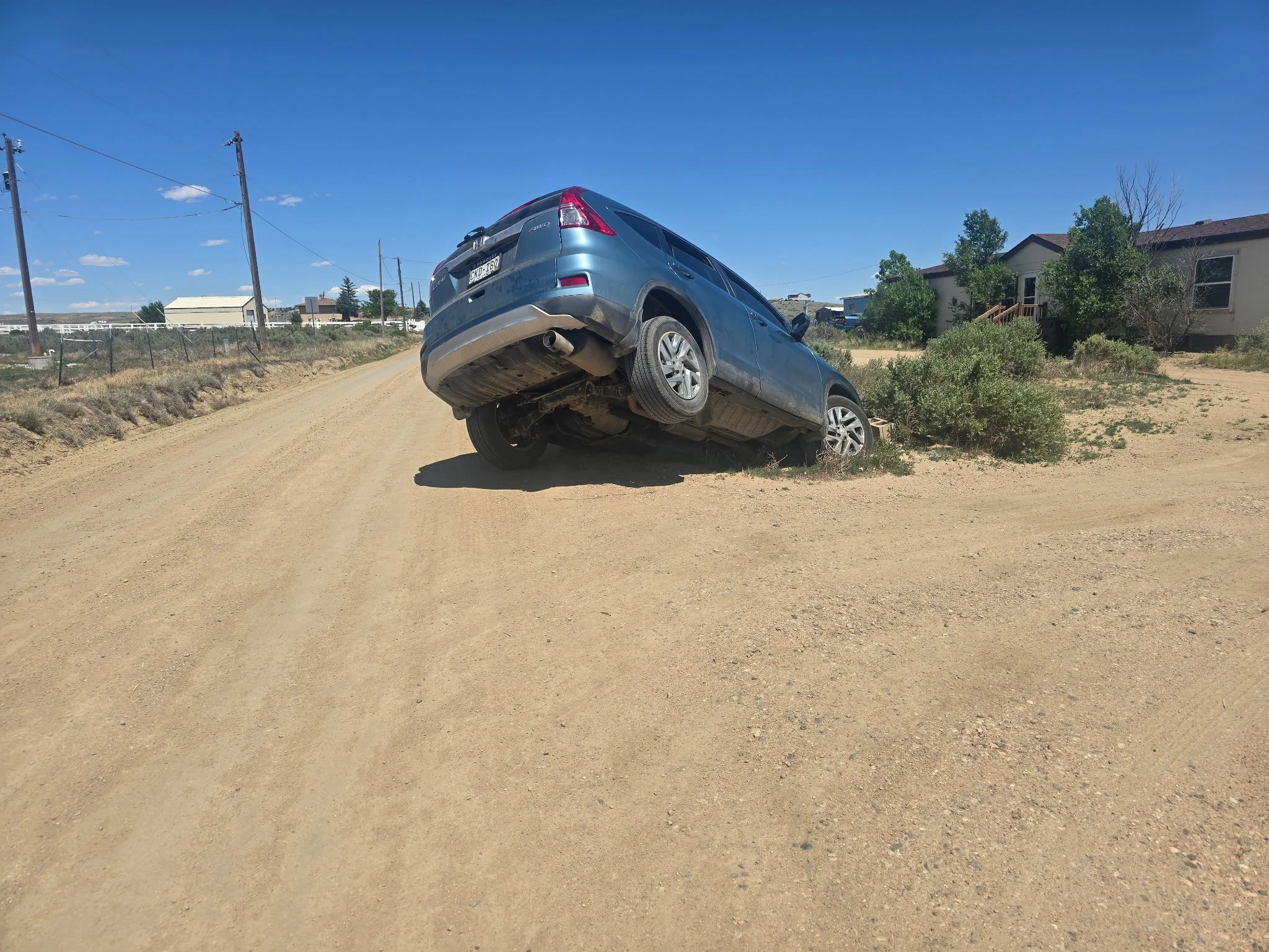 A blue SUV is tilted and hanging over the edge of a dirt road, with the rear of the vehicle slightly elevated. There is a small bush near the vehicle, and residential houses with trees in the background under a clear blue sky.