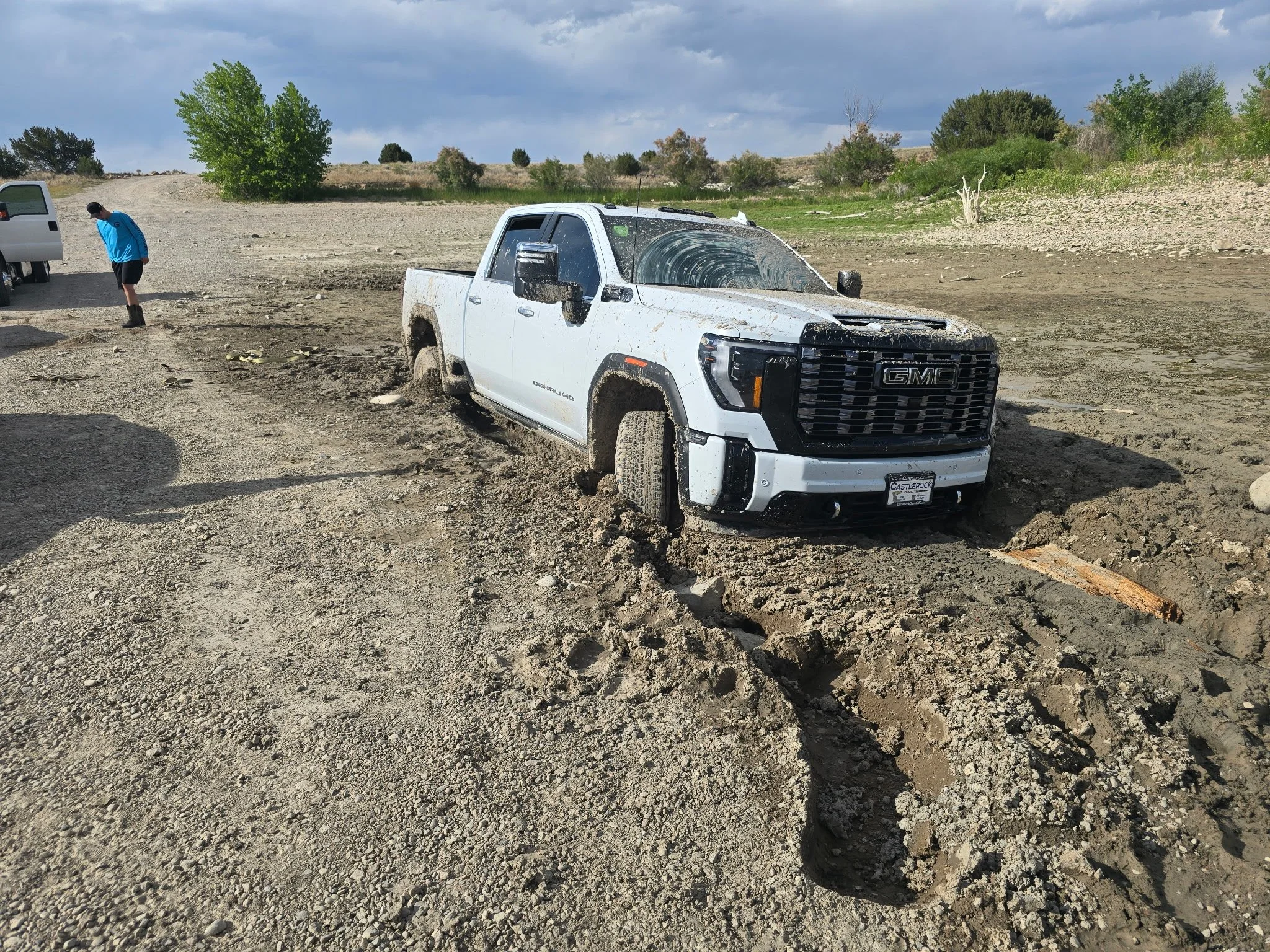 A white GMC pickup truck stuck in muddy terrain with mud splattered on its body. A person in a blue shirt and black shorts is standing near an open vehicle door, looking down at the ground. The landscape is barren with sparse trees and a cloudy sky.