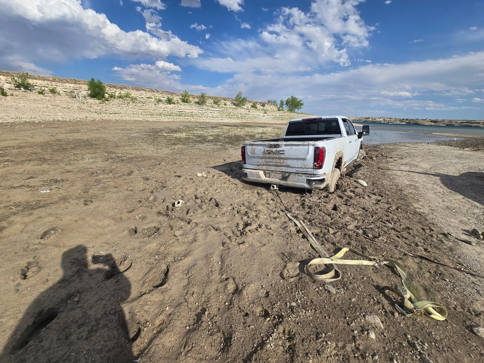 An abandoned white GMC pickup truck with mud on its tires and body, sitting on a dusty, barren ground near a body of water with a cliff and sparse greenery in the background, under a partly cloudy sky.